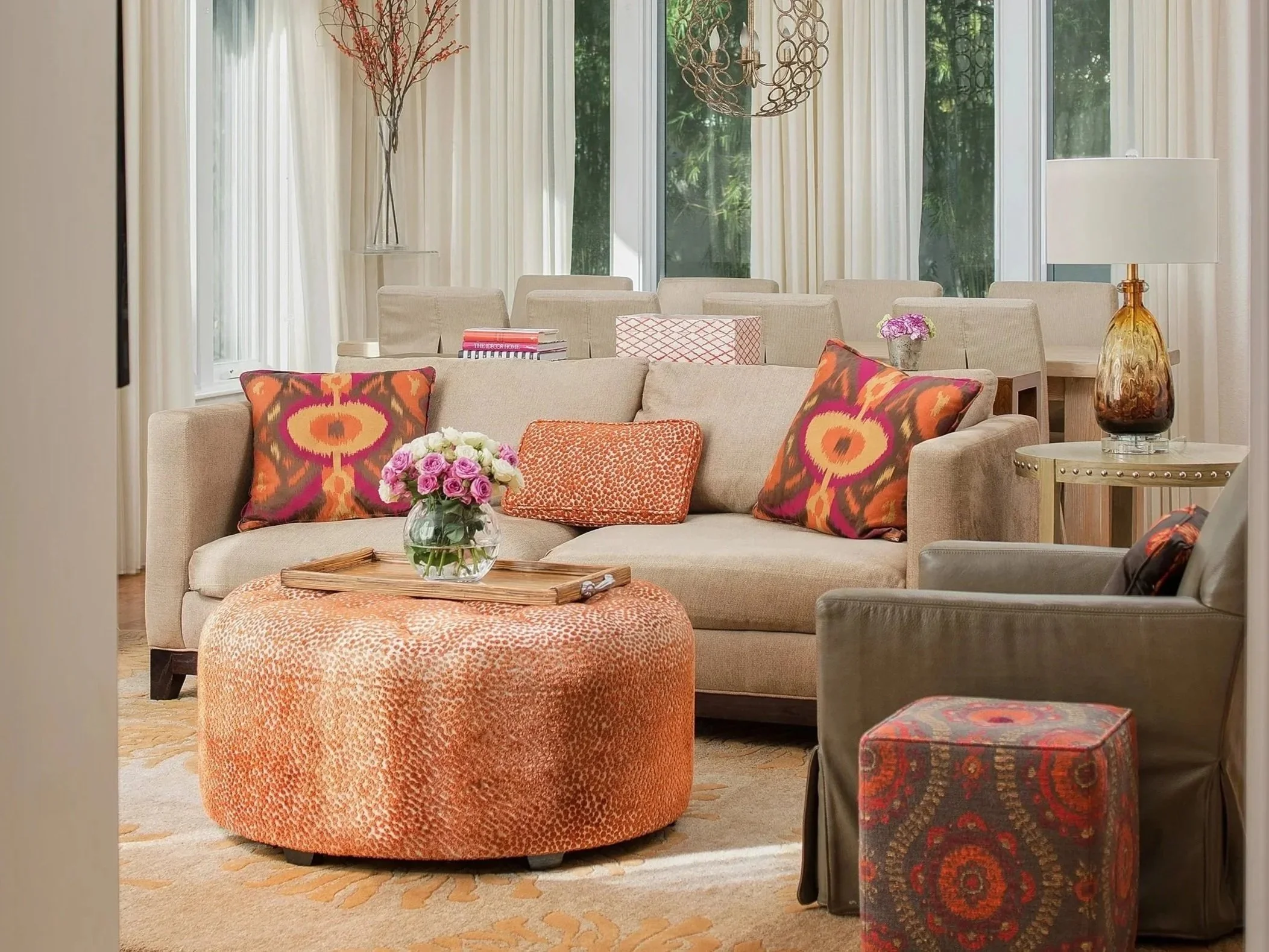 A living room with a beige sofa decorated with colorful patterned pillows, a round orange textured ottoman with a wooden tray and flower vase, a gray armchair, a side table with a decorative glass lamp, and a dining area in the background with a table, chairs, and a chandelier.