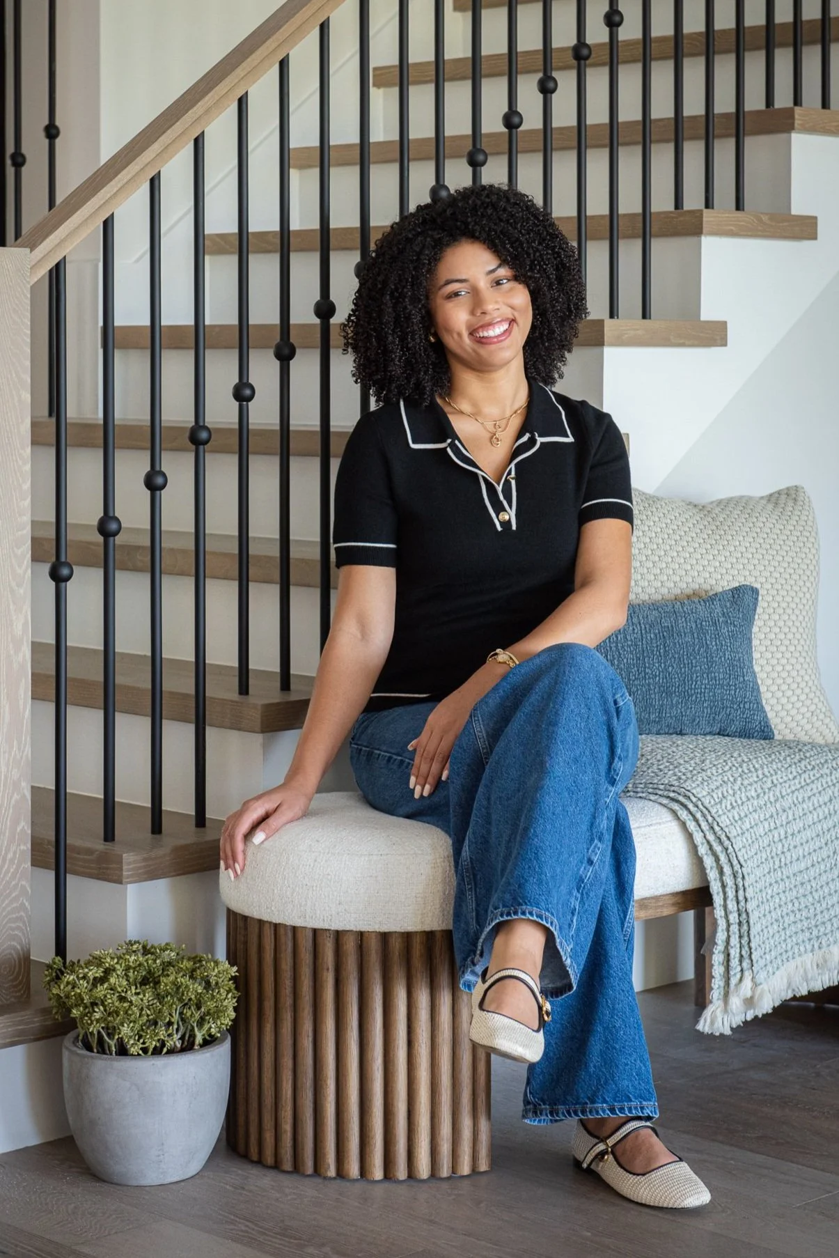 A smiling woman with curly hair sitting on a rounded upholstered bench in front of a staircase with wooden steps and black metal railings, next to a potted plant and a cozy blanket on a woven bench.
