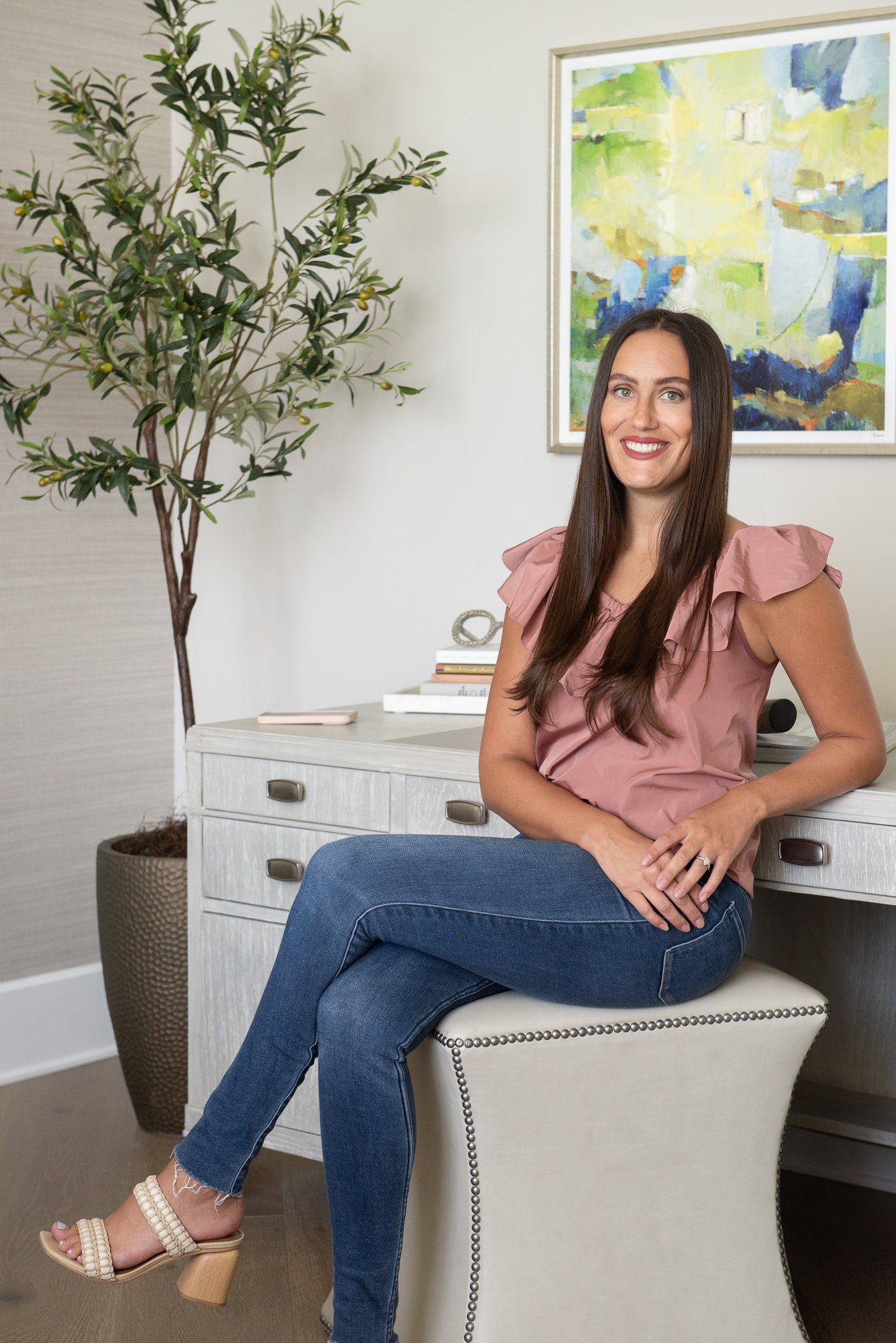 A woman with long brown hair, wearing a pink ruffled blouse, blue jeans, and beige open-toe heeled sandals, is sitting on a cream-colored ottoman, smiling, in a room with a large potted plant and colorful abstract painting on the wall behind her.