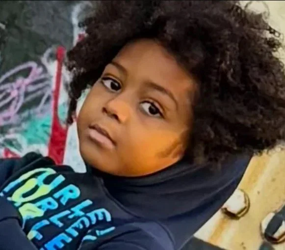 Young girl with curly hair and a serious expression, wearing a dark hoodie, standing outdoors near a graffiti-covered wall.