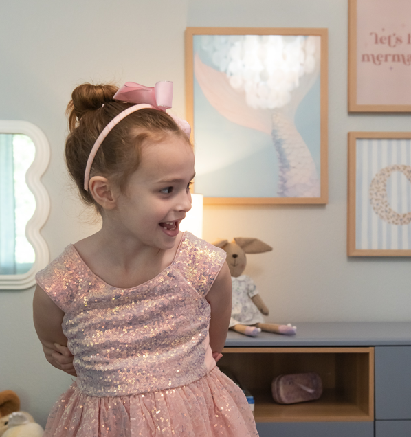 A young girl with red hair tied in a bun, wearing a pink sparkly dress and a pink bow headband, smiling and looking to her right inside a decorated room.