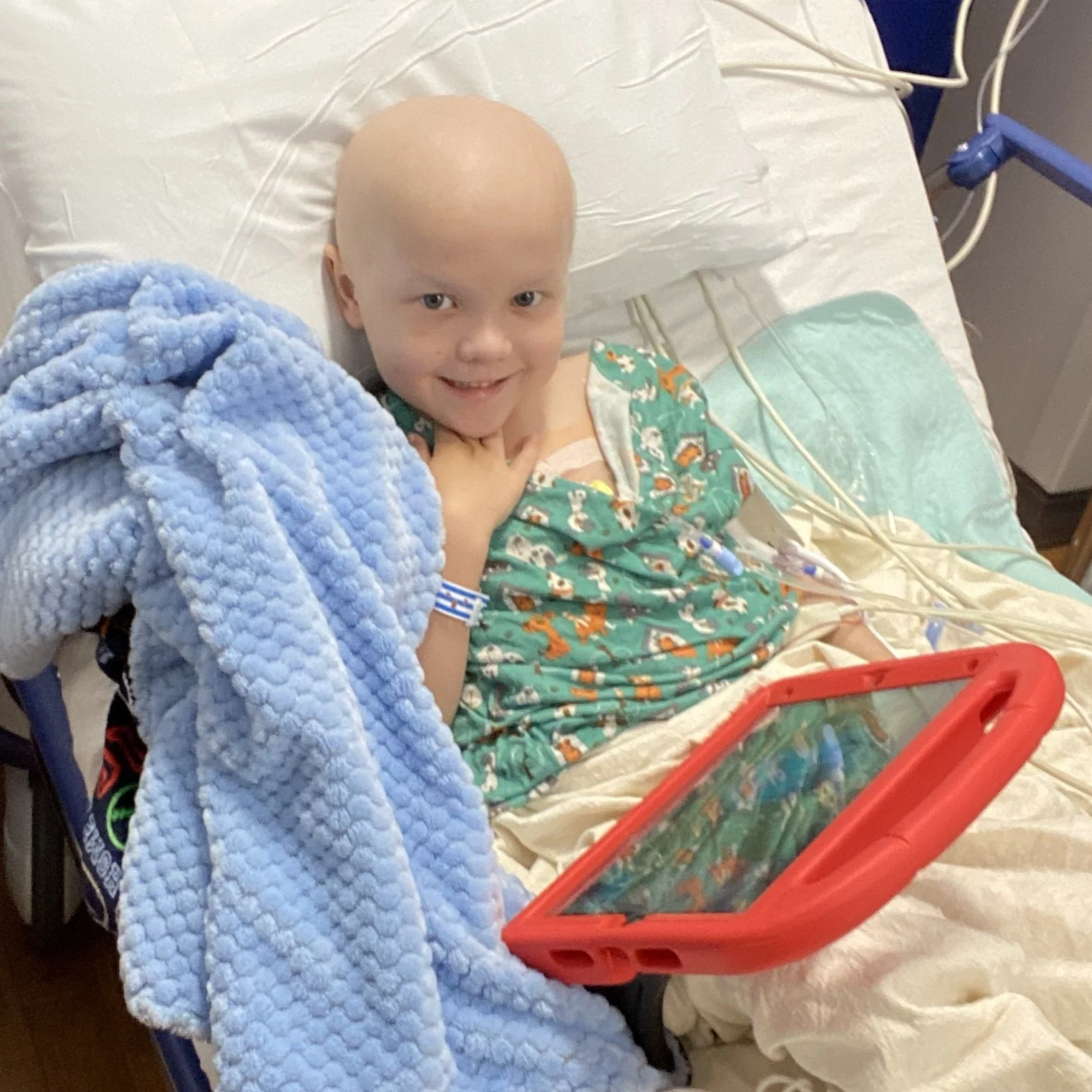 A young boy in a hospital bed, smiling, with a shaved head, wearing a hospital gown with colorful patterns. He has a hospital bracelet on his wrist and is holding a red tablet. There are medical tubes attached to him, and a blue textured blanket partially covers him.