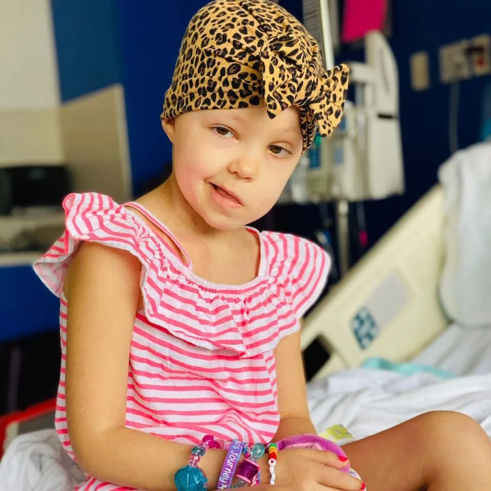 A young girl wearing a leopard print headscarf and a pink and white striped dress sitting on a hospital bed.