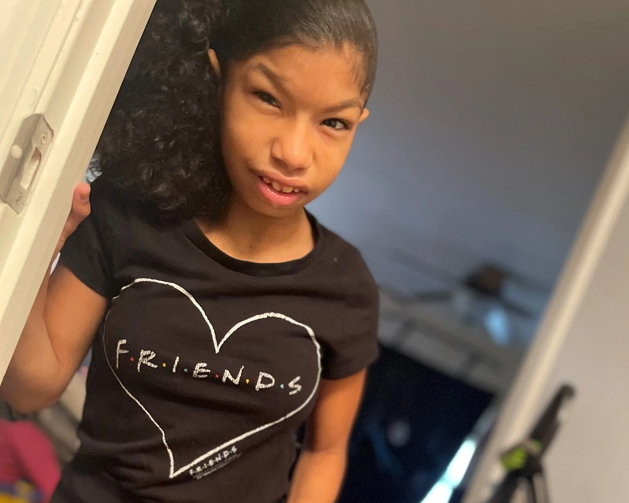 A young girl with curly hair peeking around a doorframe, wearing a black T-shirt with the word 'FRIENDS' inside a heart illustration.