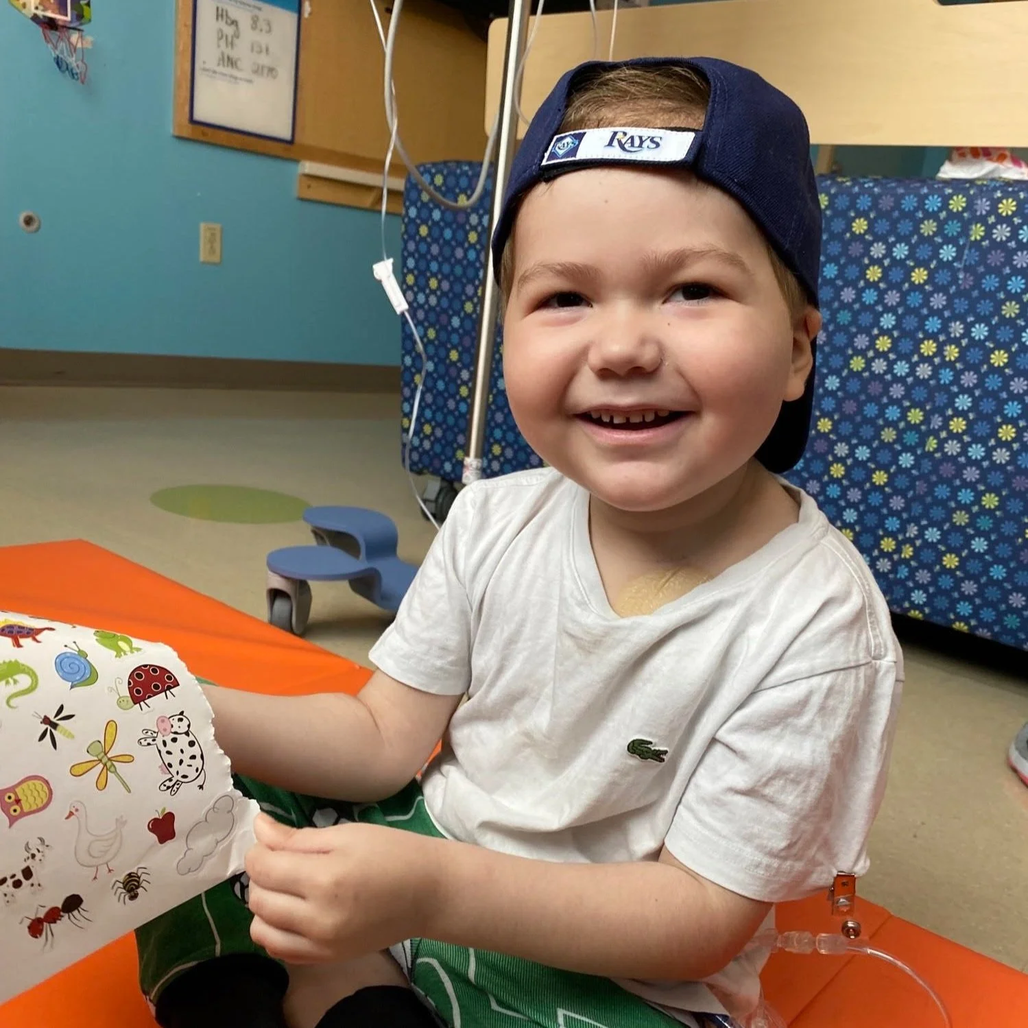 A young boy with a happy expression, wearing a navy cap backwards and a white t-shirt, sitting on an orange mat in a hospital or medical setting. He has a medical tube connected to his arm and a bandage on his chest. There are medical equipment and colorful furniture in the background.