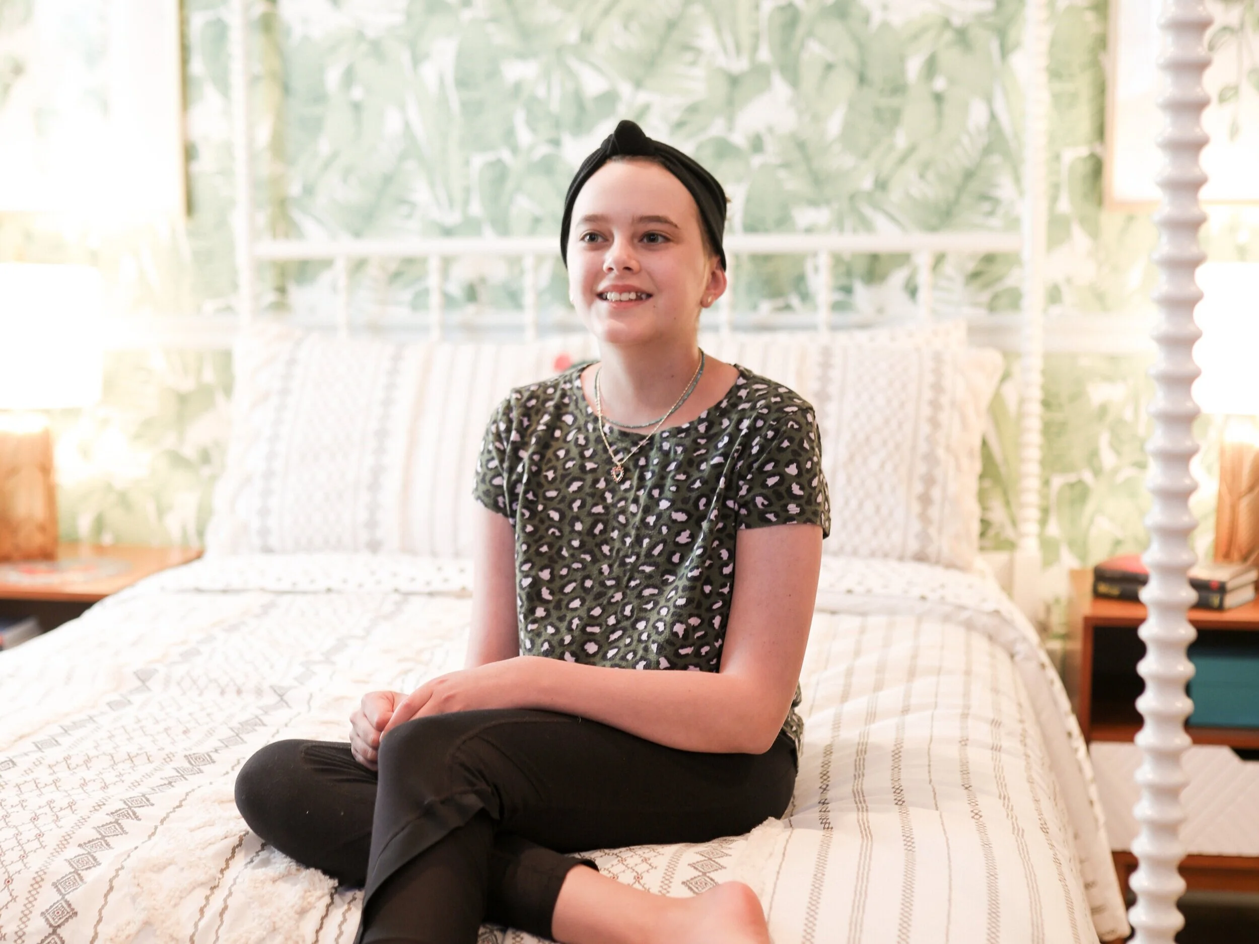 A young girl with short dark hair, wearing a black headband, sitting cross-legged on a bed in a bedroom with green patterned wallpaper and white bedding, smiling and looking to the side.