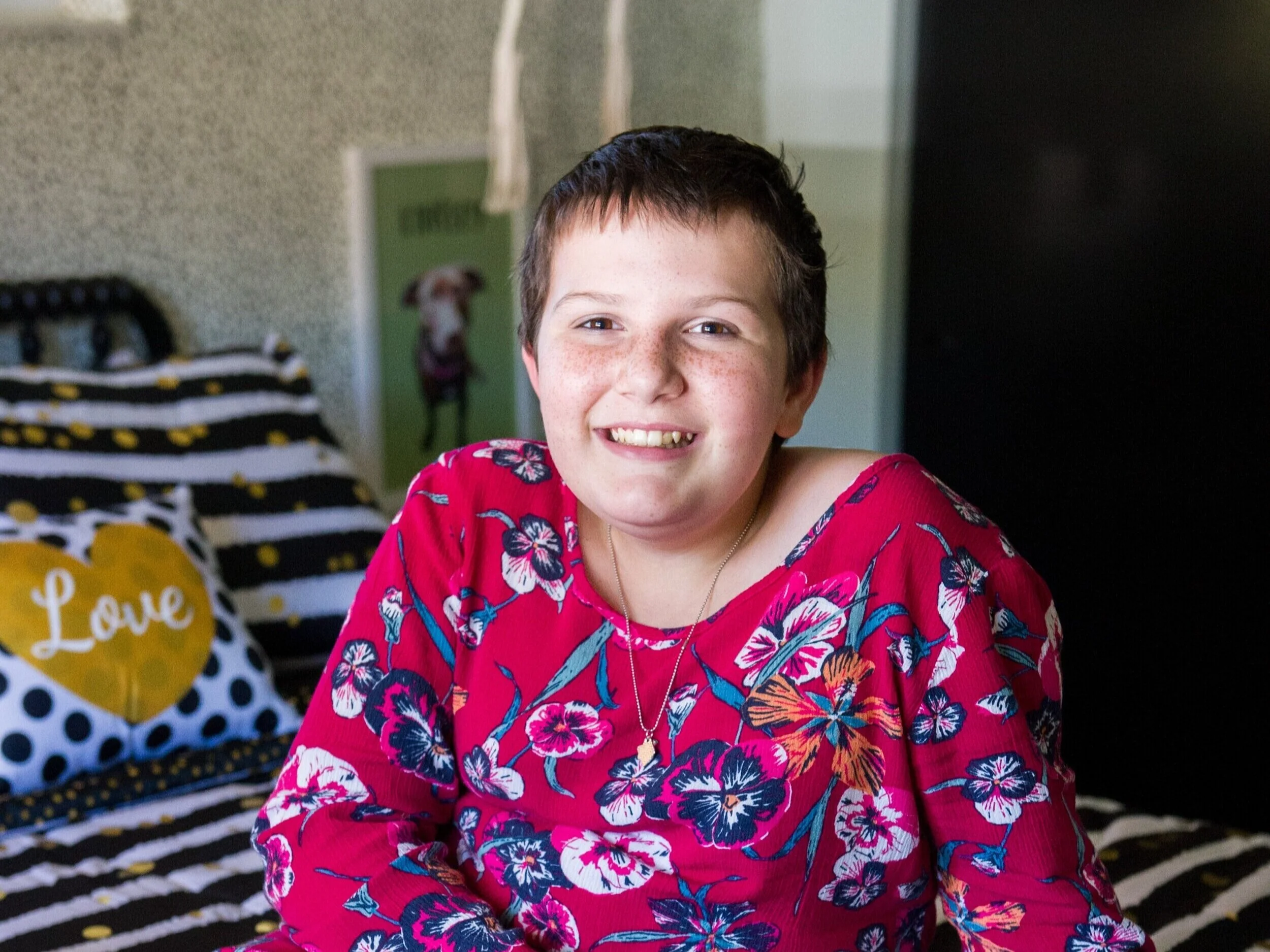 Young girl with short brown hair, smiling, wearing a pink floral shirt, sitting on a bed with black and white bedding that has yellow accents and a pillow that says 'Love' in white letters within a yellow heart.