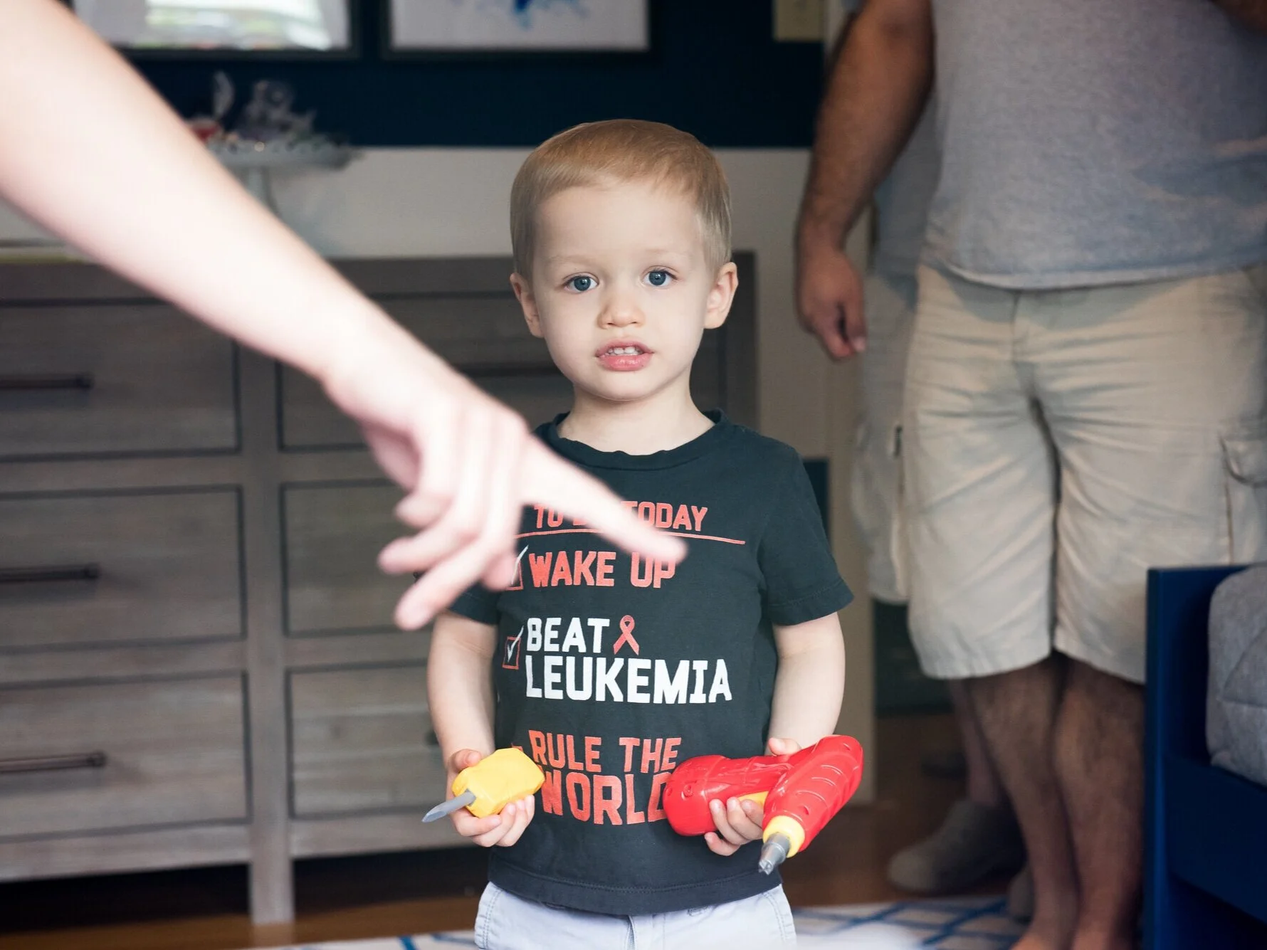 A young boy with blonde hair and blue eyes stands indoors, holding a yellow and gray toy knife in one hand and a red toy drill in the other. He is wearing a black T-shirt with red and white text supporting leukemia awareness. A person in a gray T-shirt and beige shorts is visible in the background.