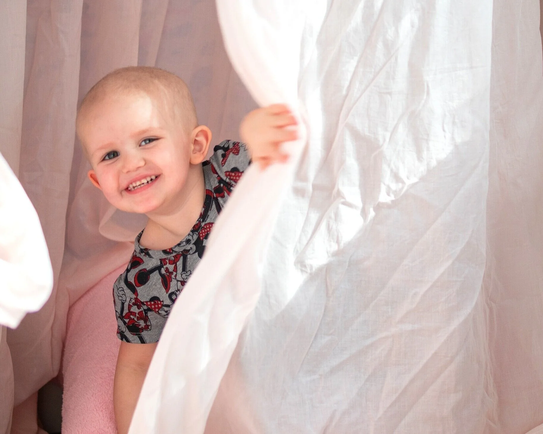 A young smiling boy with short blonde hair and a gray shirt with Minnie Mouse print, peeking through white curtains.