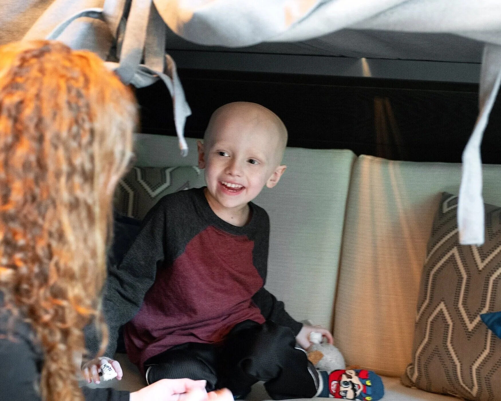 A young boy with a shaved head smiling while sitting on a couch, talking to a person with curly red hair.