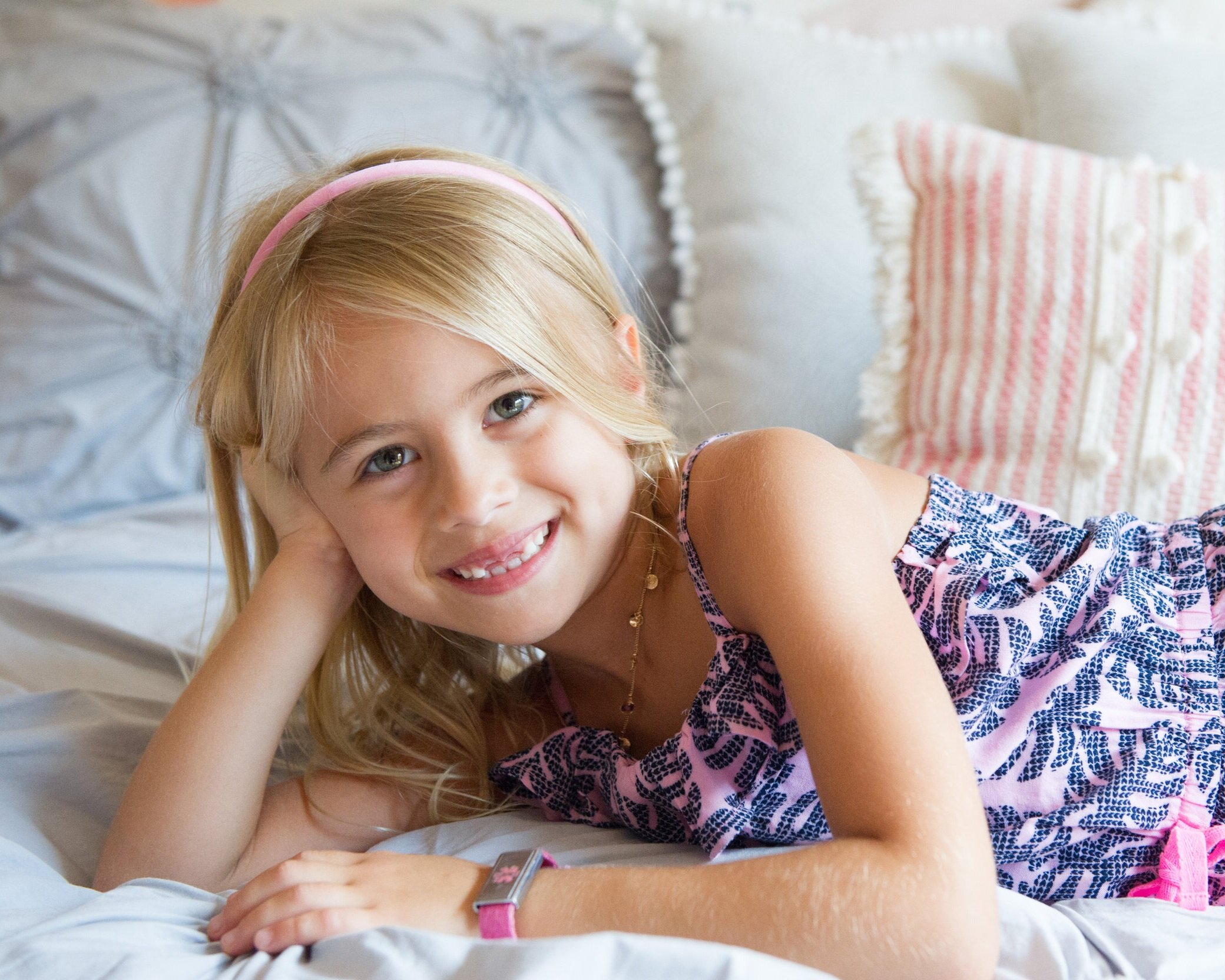 A young girl with blonde hair lying on a bed, smiling at the camera. She is wearing a pink headband, a purple patterned dress, and a pink watch, with pillows in the background.