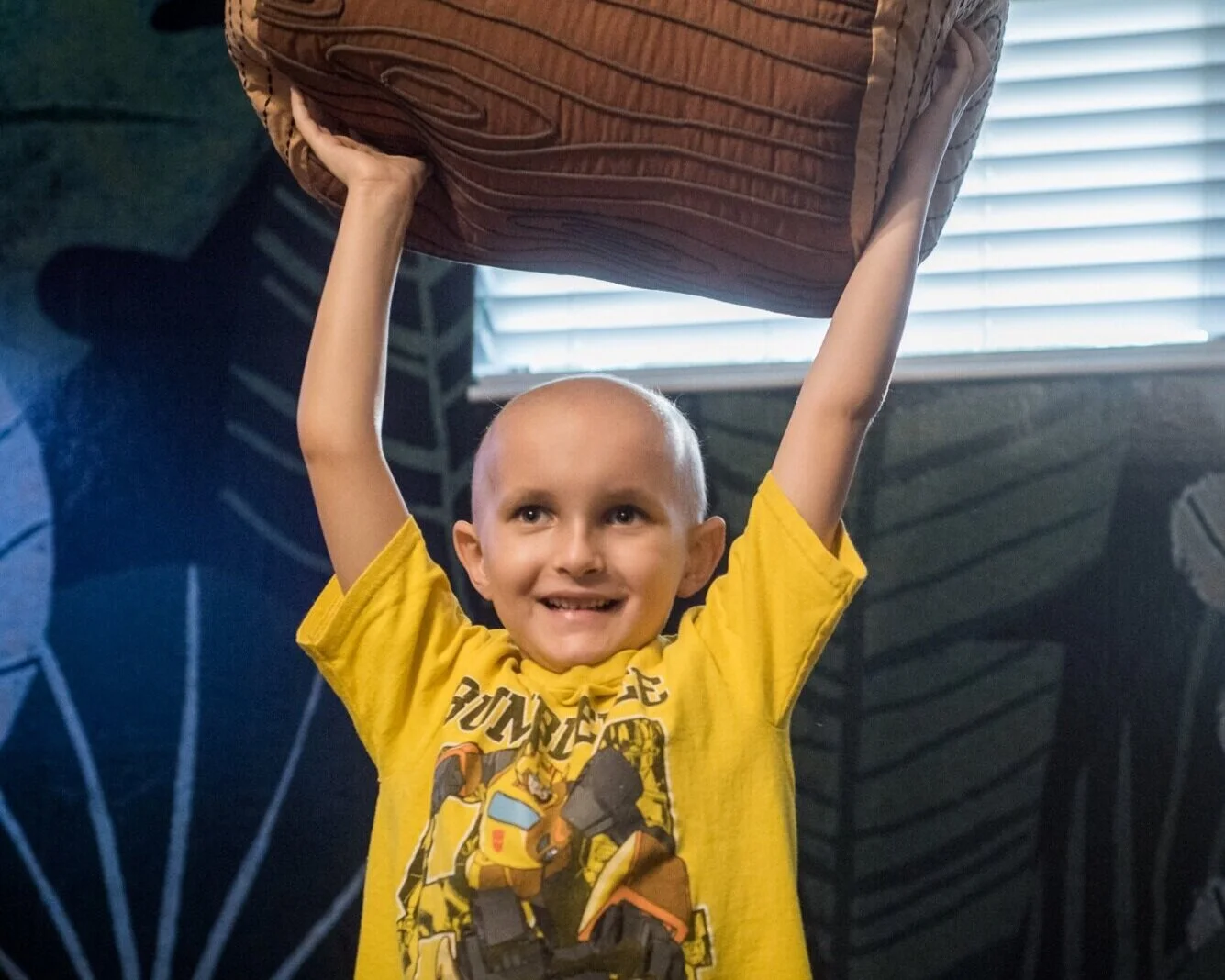 A young boy with a shaved head, wearing a yellow T-shirt with a graphic, is holding a large brown cushion overhead with both hands and smiling.