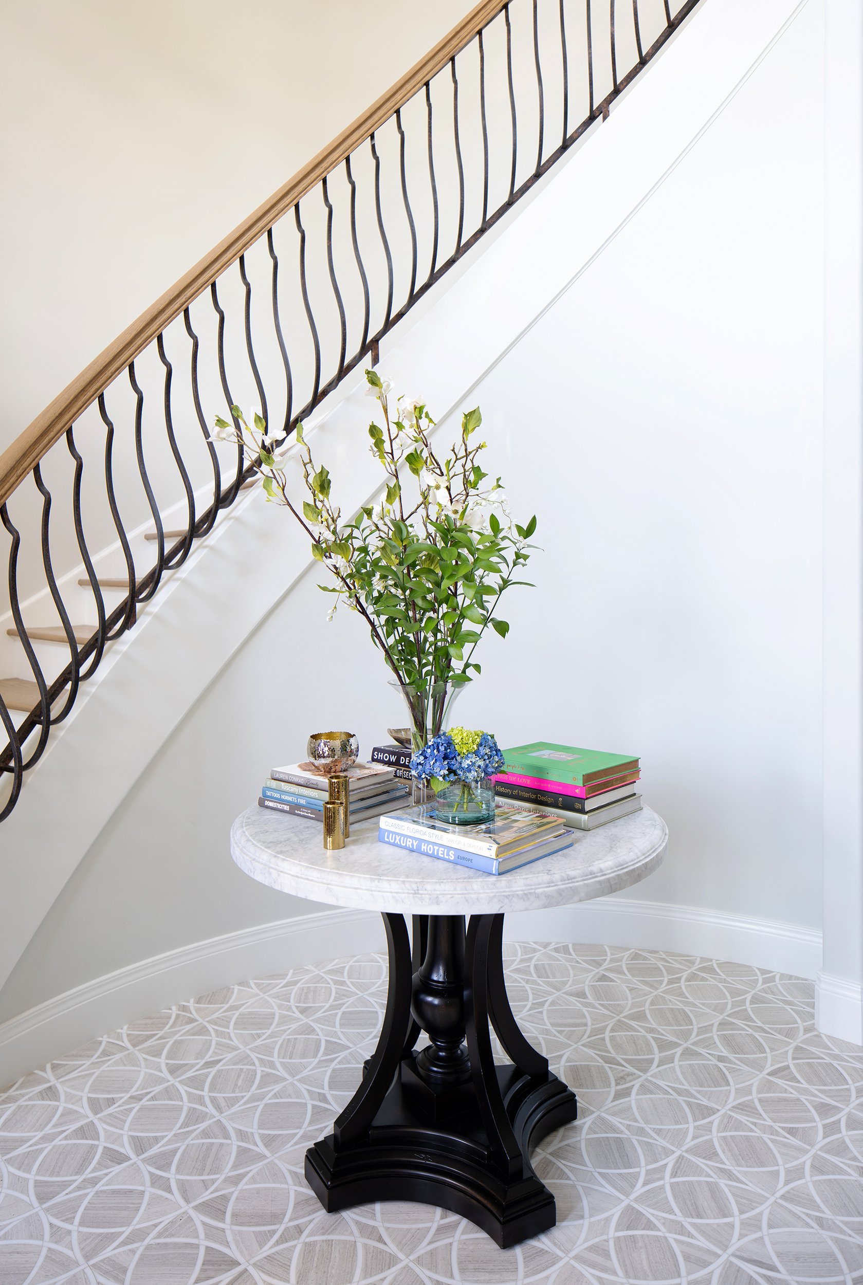 Decorative round table with a marble top, black base, on a patterned carpet, holding books, a vase of greenery and flowers, and small gold decorative objects, next to a staircase with a wooden handrail and black wrought iron balustrade.