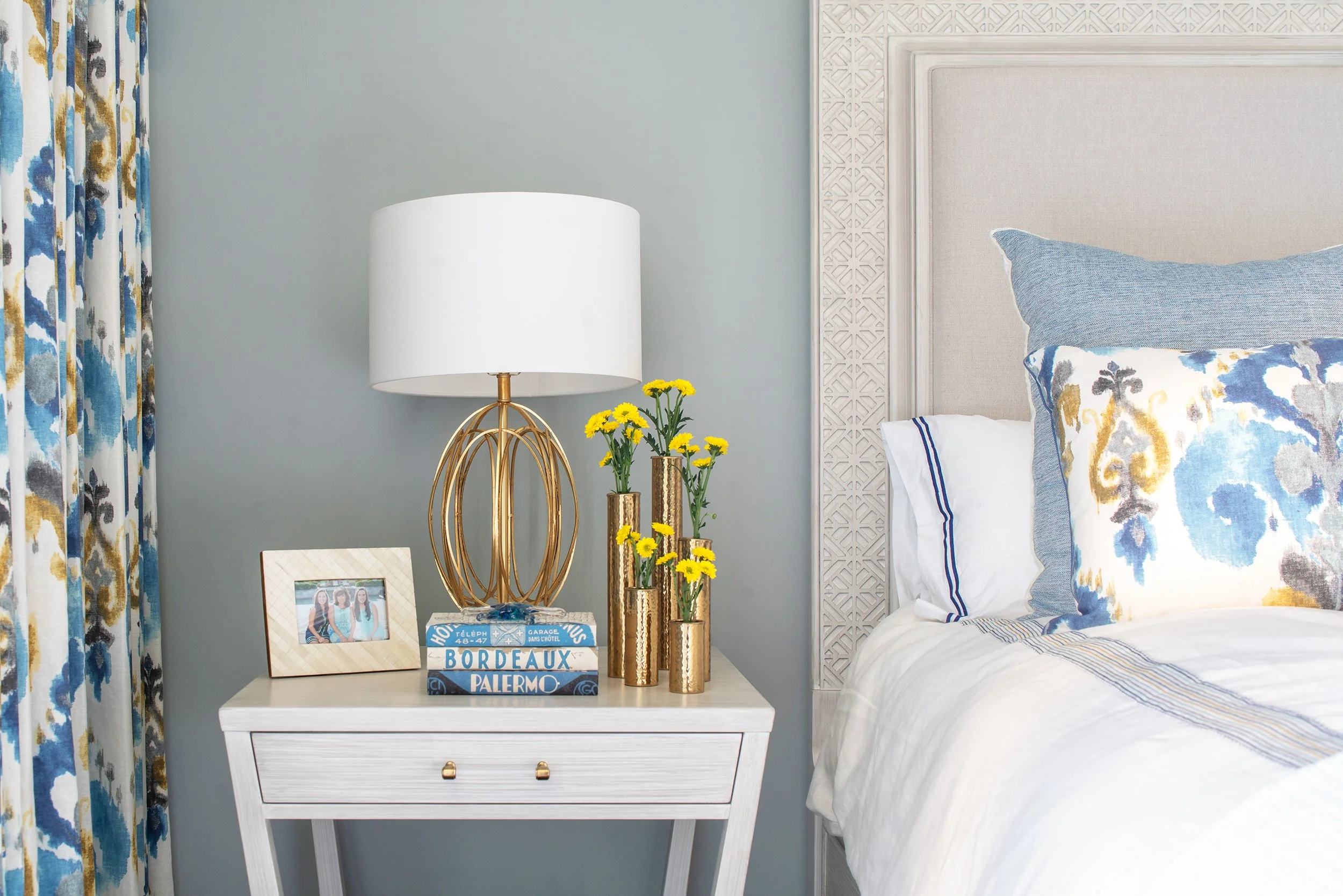 Close-up of a white bedside table with a gold lamp, a framed photo, and three gold vases with yellow flowers, next to a bed with a decorative blue and white pillow and a large headboard, in a bedroom with light blue walls and patterned curtains.