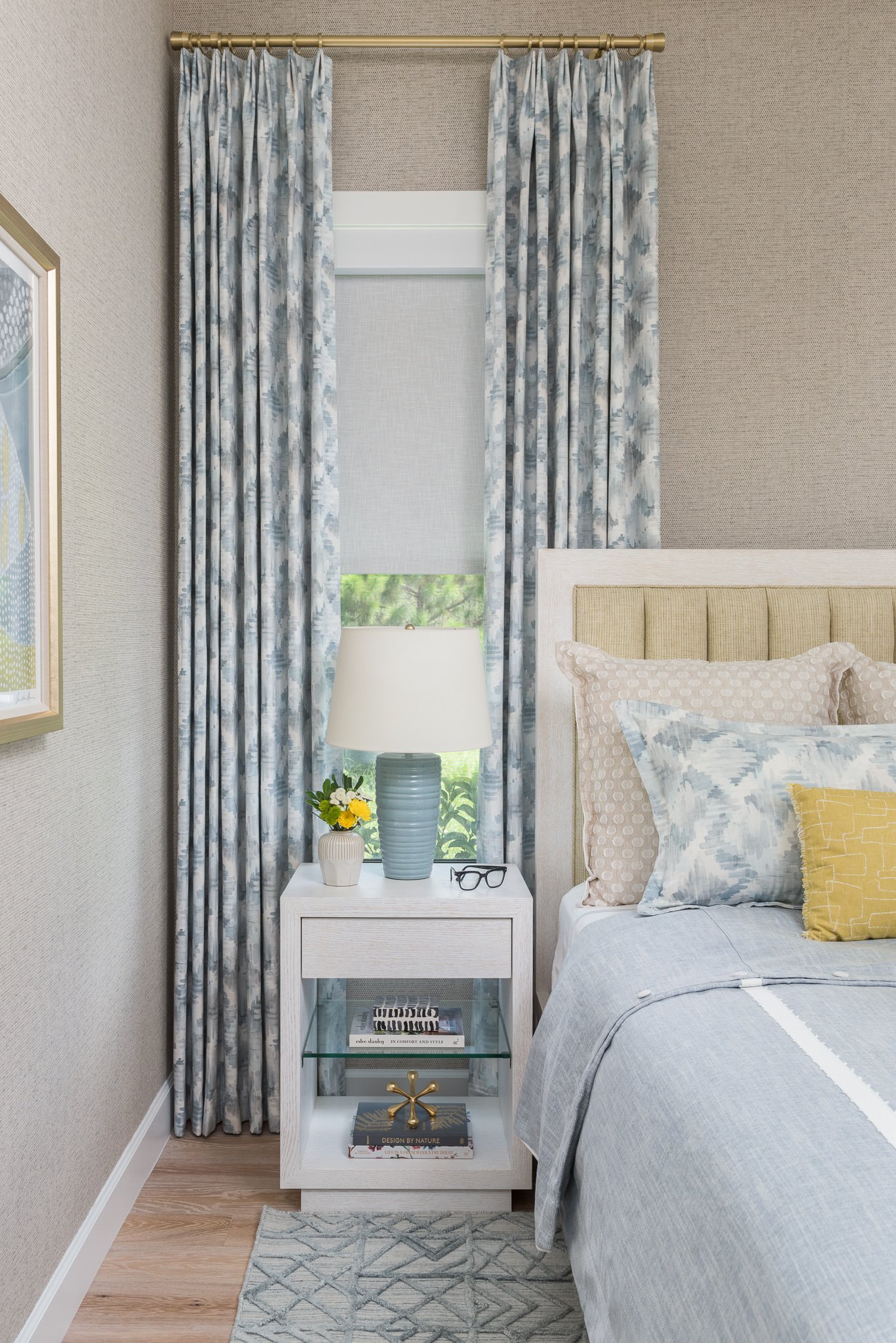 A bedroom with a window dressed in blue patterned curtains and a white roller shade. There is a white nightstand with a blue ceramic lamp, a vase with yellow flowers, and some books with a pair of glasses on top. Part of a bed with a beige and blue b
