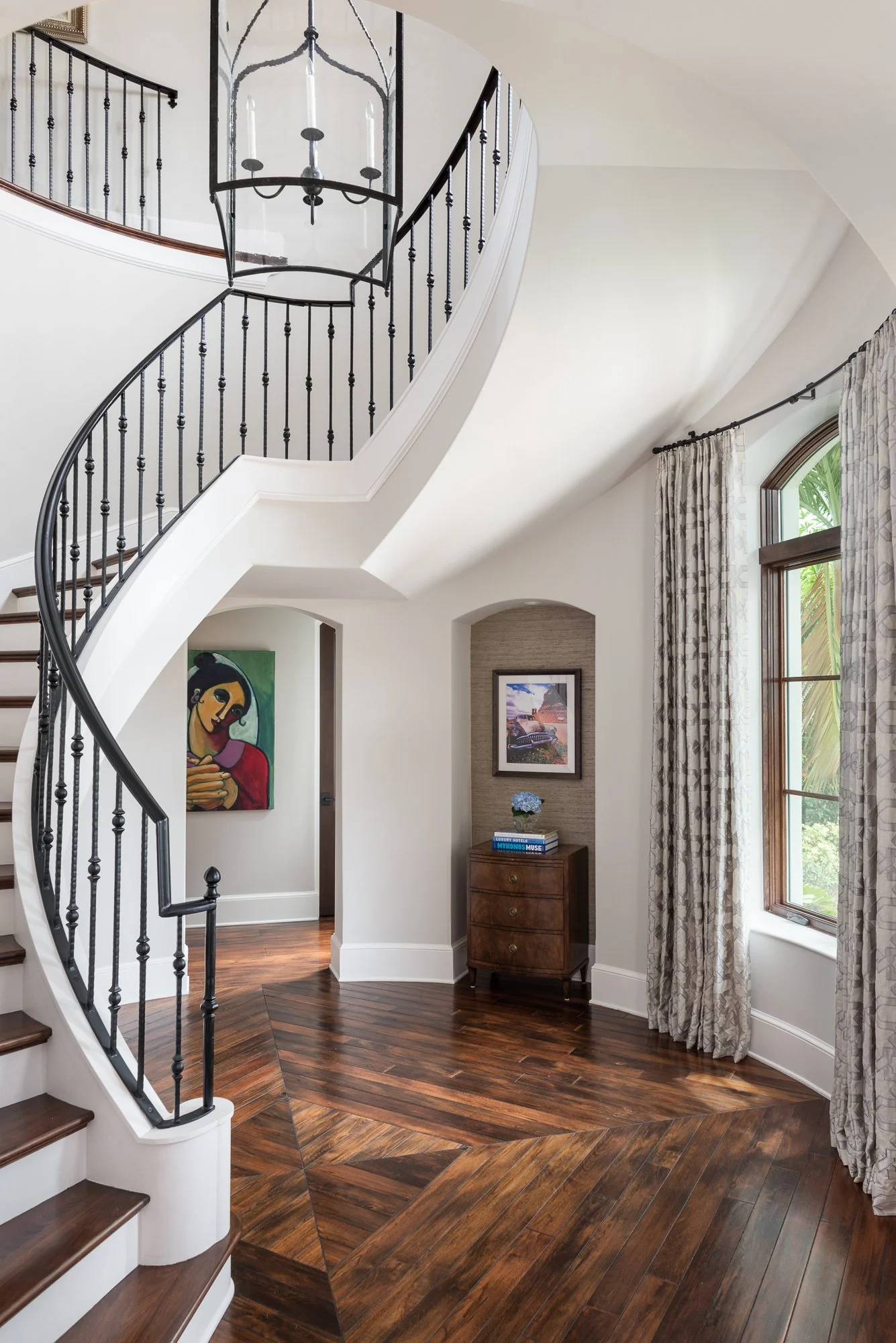 Interior view of a home with a curved staircase, large window with patterned curtains, wooden floor, and artwork on the walls.