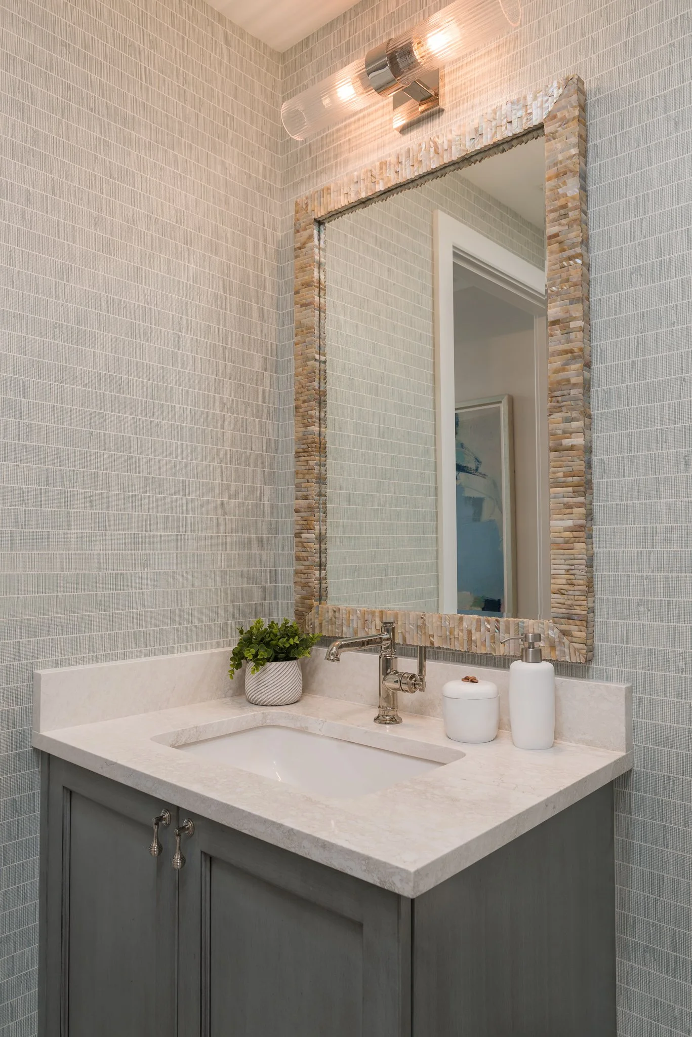 Bathroom vanity with a large mirror, light-colored tile walls, a small potted plant, a soap dispenser, and a cabinet below.