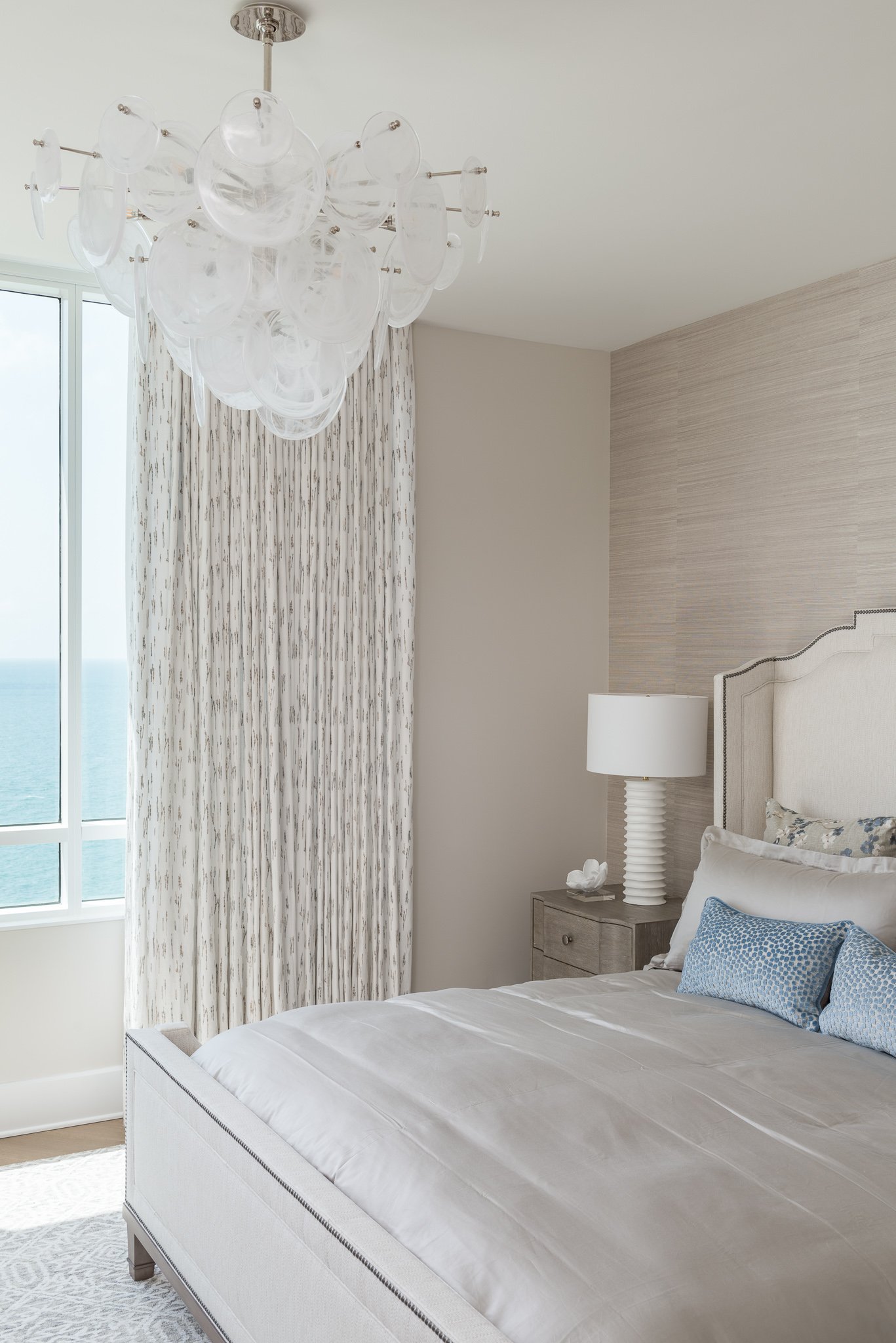 A bedroom with a large glass window, a chandelier with clear glass orbs, a bed with white sheets and blue pillows, a nightstand with a white lamp and decor, and patterned curtains, overlooking an ocean.