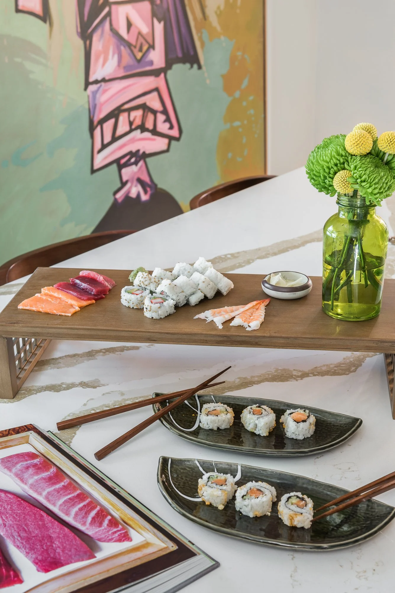Sushi assortment on a two-tiered display with chopsticks, a small dish of wasabi, and a vase of green and yellow flowers, set on a white table with a colorful abstract painting in the background.