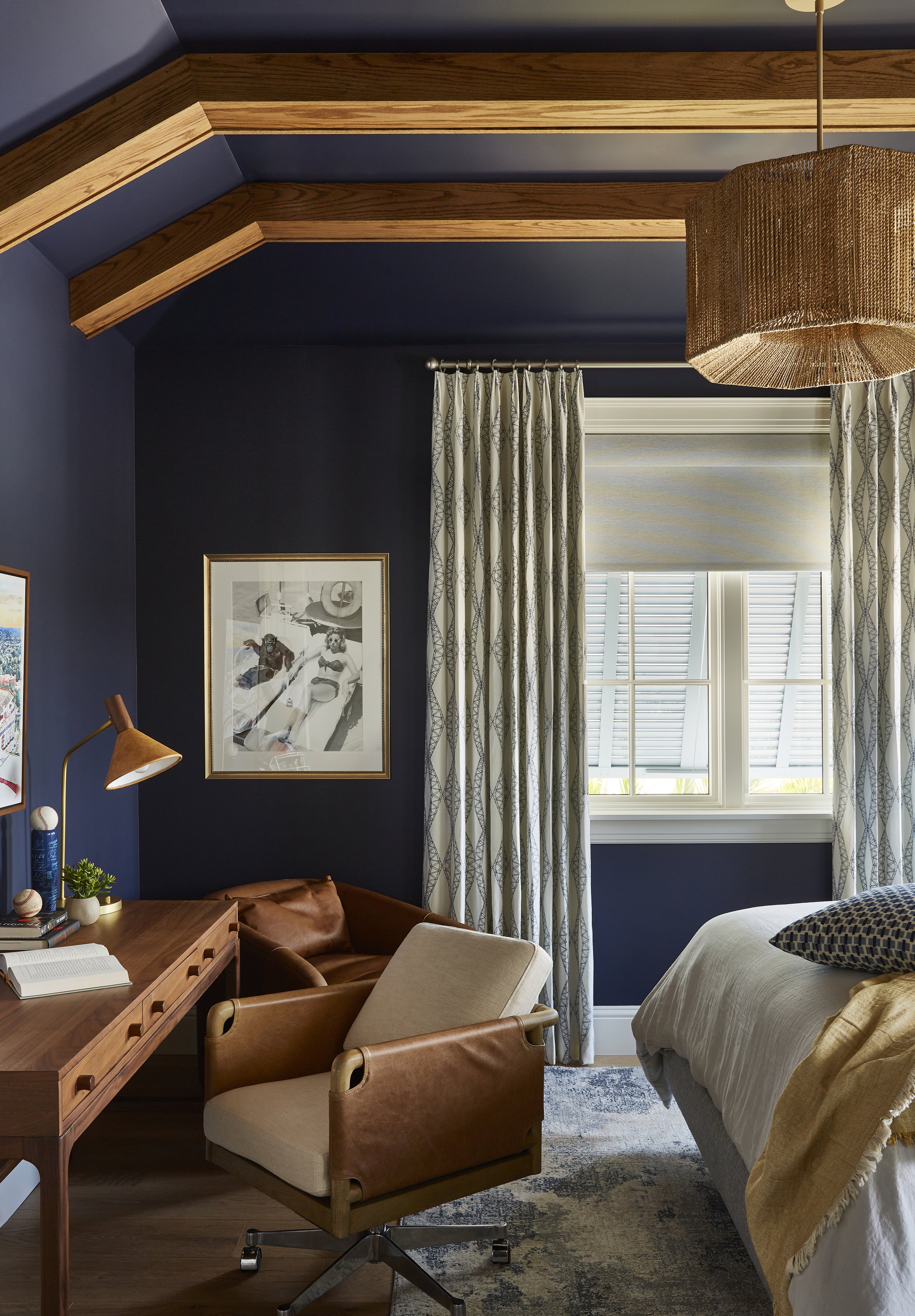 A cozy bedroom with navy blue walls, a large window with white shutters, tropical patterned curtains, a bed with white bedding and dark patterned pillows, a wooden desk with a beige and leather chair, a brown leather armchair, and wall art. A large w