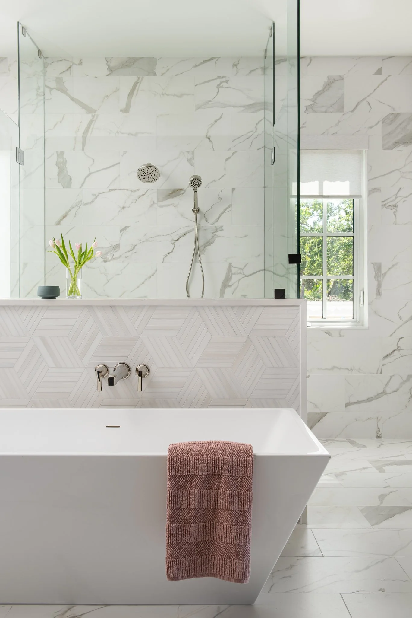 Modern bathroom with a white bathtub, pink towel, and marble tile walls. The shower area has two showerheads and a window with view of trees.