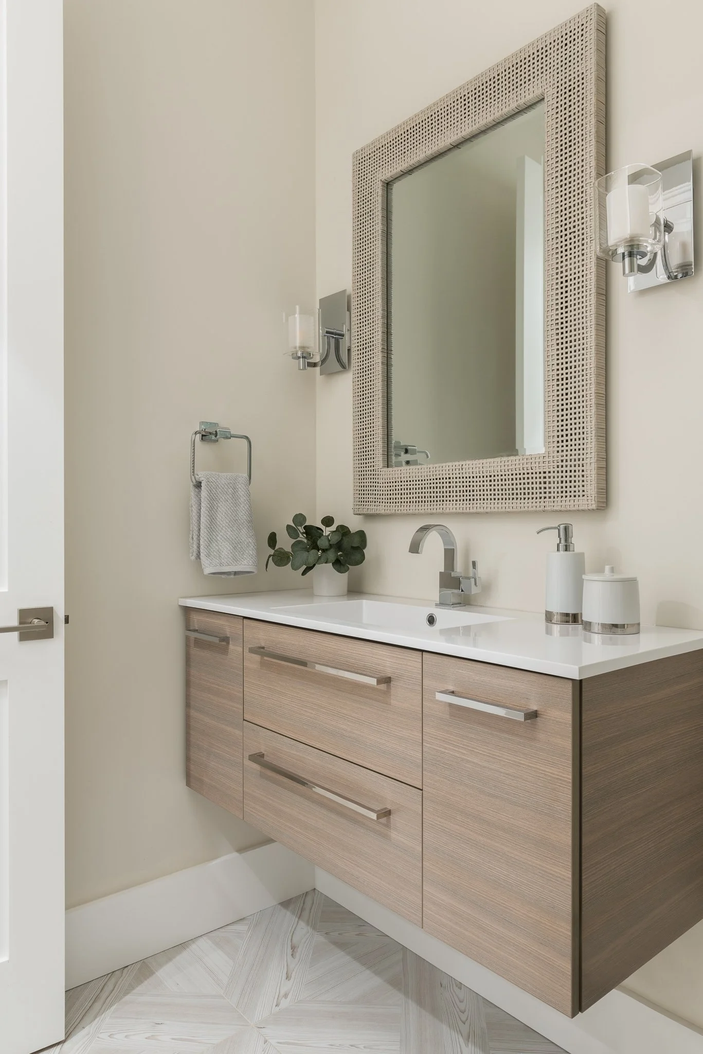 Modern bathroom vanity with a wood finish, white countertop, large framed mirror, wall-mounted light fixtures, a faucet, soap dispenser, and a small potted plant. Towel hanging on a wall-mounted holder.