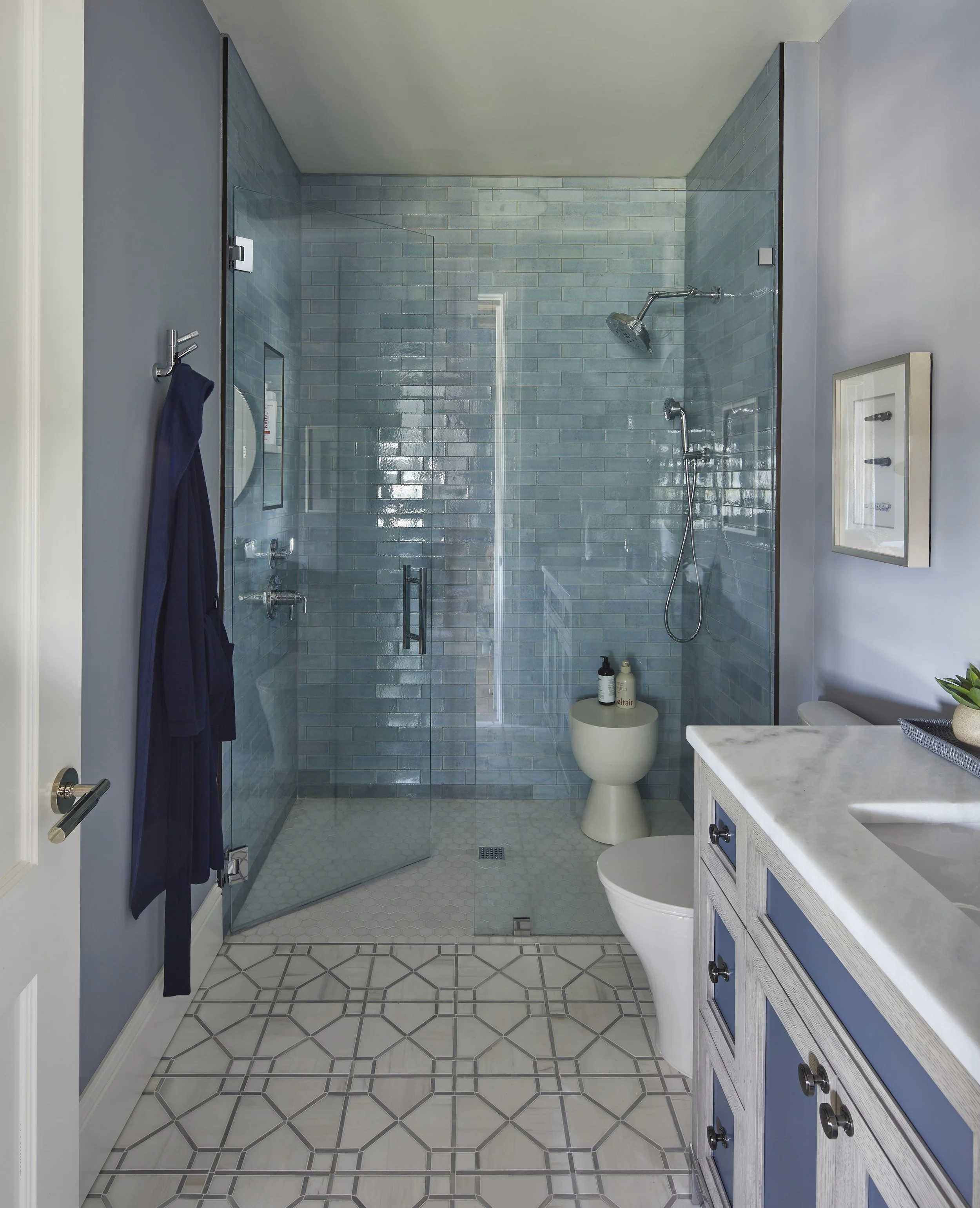 Modern bathroom with blue tiled walk-in shower, white floor tiles with a geometric pattern, a white vanity with a marble countertop, and a small plant on the right side.