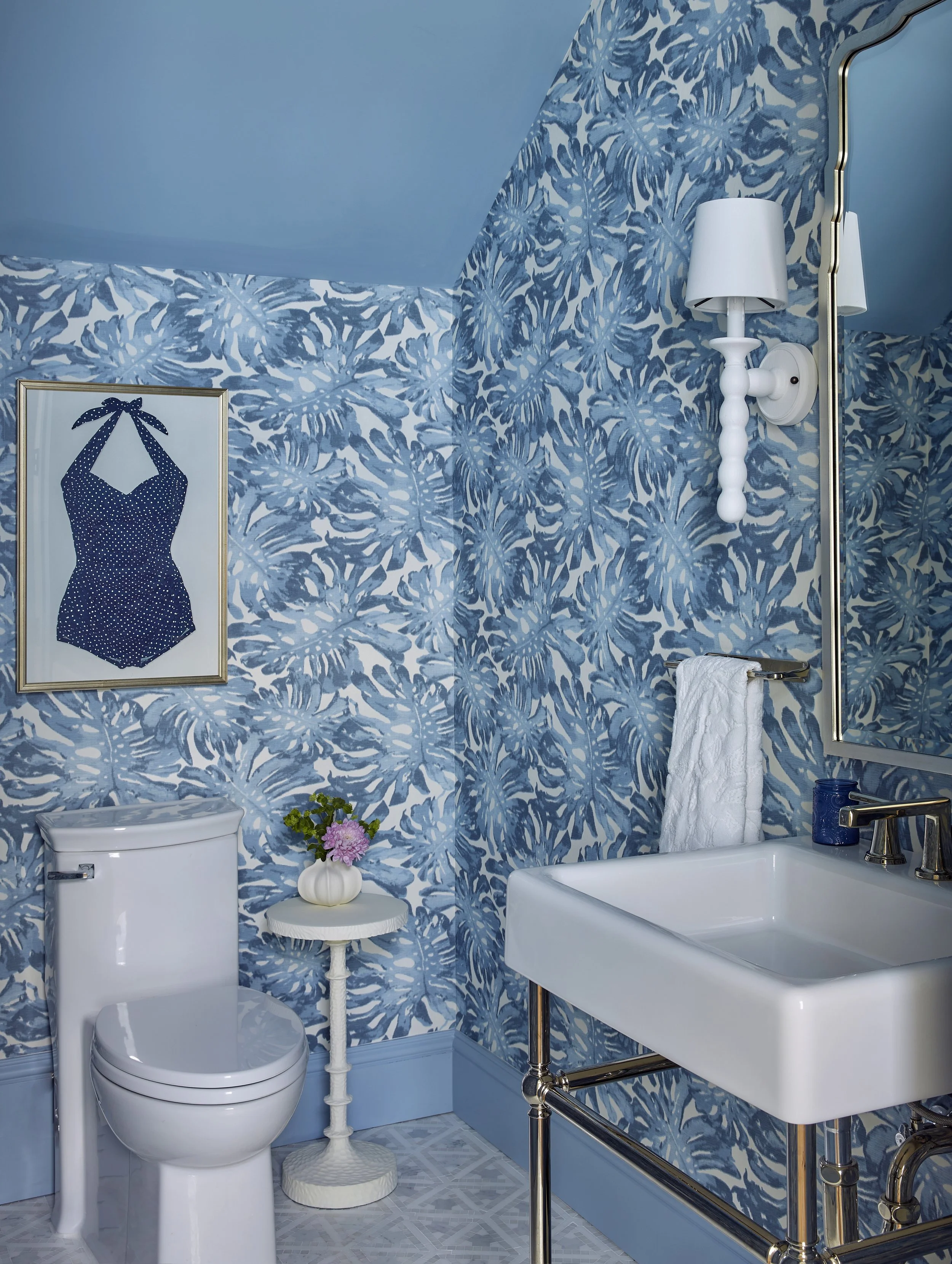 A bathroom with blue and white patterned wallpaper, a framed art of a polka-dotted swimsuit, a white porcelain toilet, a small white side table with a vase of flowers, a white towel hanging on a rail, a white rectangular sink with brass fixtures, a w