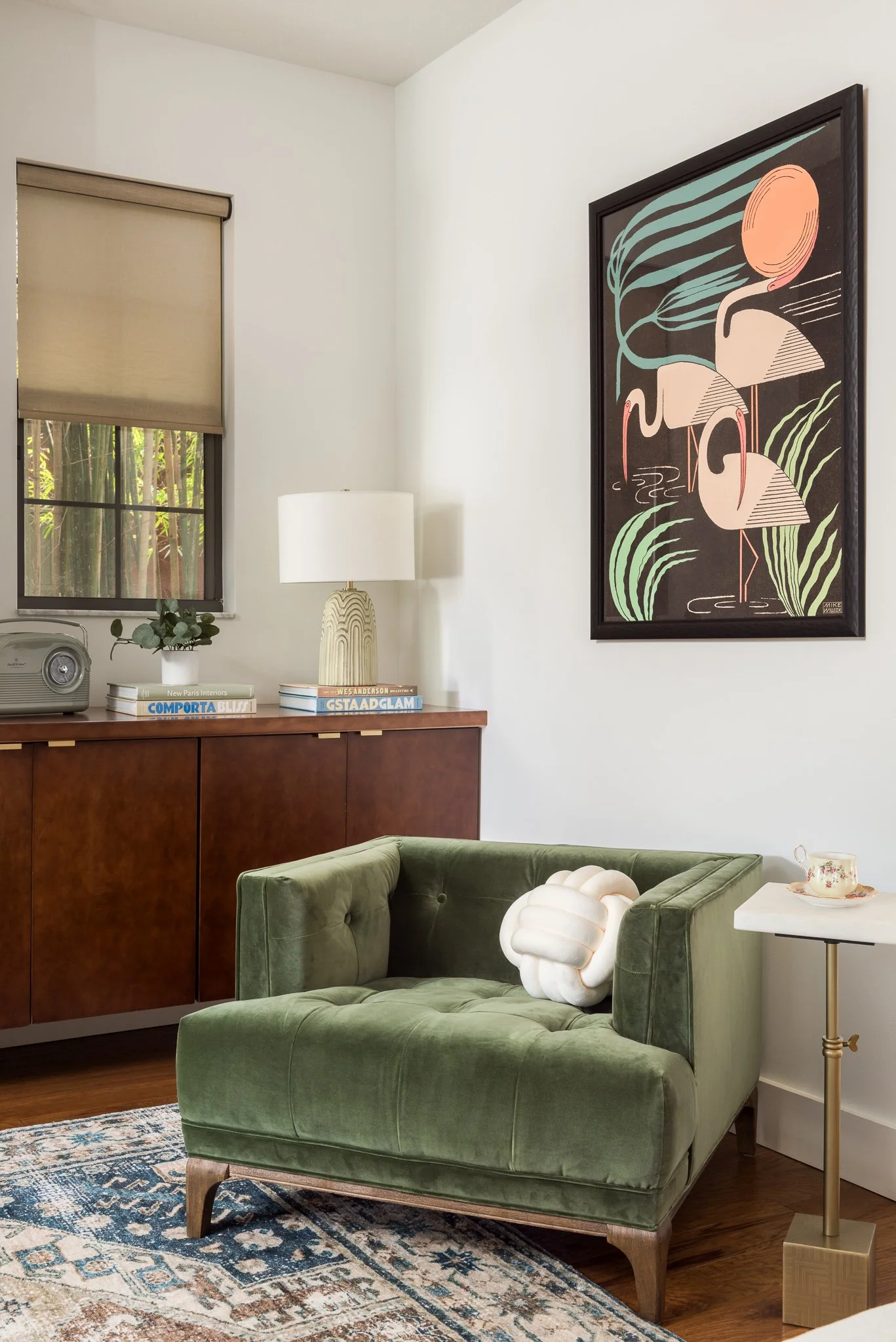 A cozy living room corner with a green velvet loveseat, a side table with a teacup, a wooden cabinet with books and a lamp, a window with a beige roller shade, a decorative pillow shaped like a brain on the loveseat, a framed artwork of flamingos on 