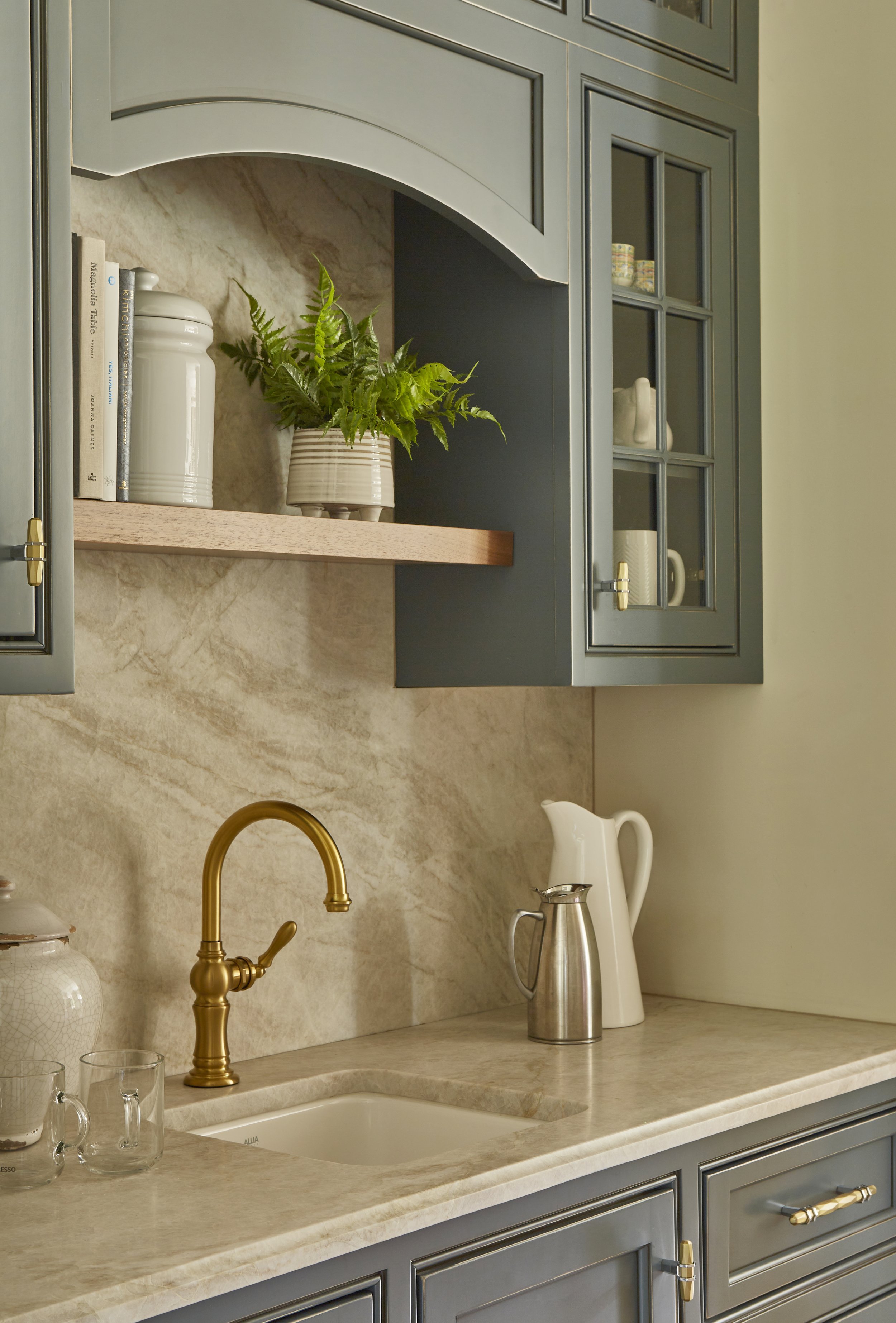 A kitchen countertop with a gold faucet, white and silver pitchers, a beige marble backsplash, and gray cabinets with glass doors and gold handles. A potted plant and some books are on an open shelf above.