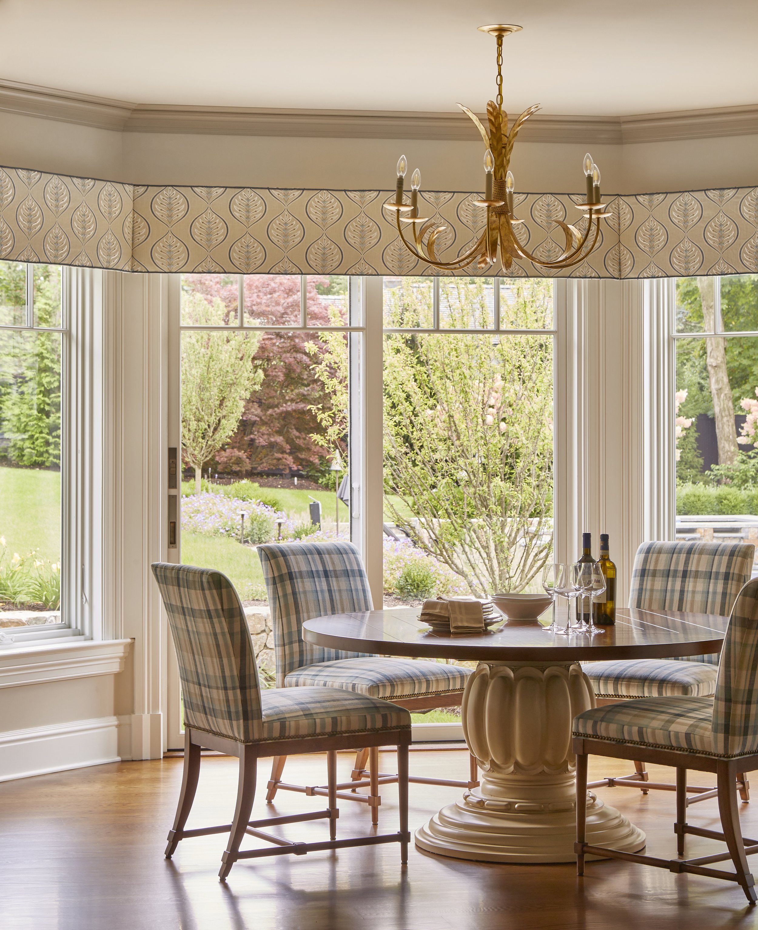 Dining room with a round wooden table and four plaid upholstered chairs, large windows showing a garden with trees and flowers, a chandelier hanging from the ceiling, and a wooden floor.