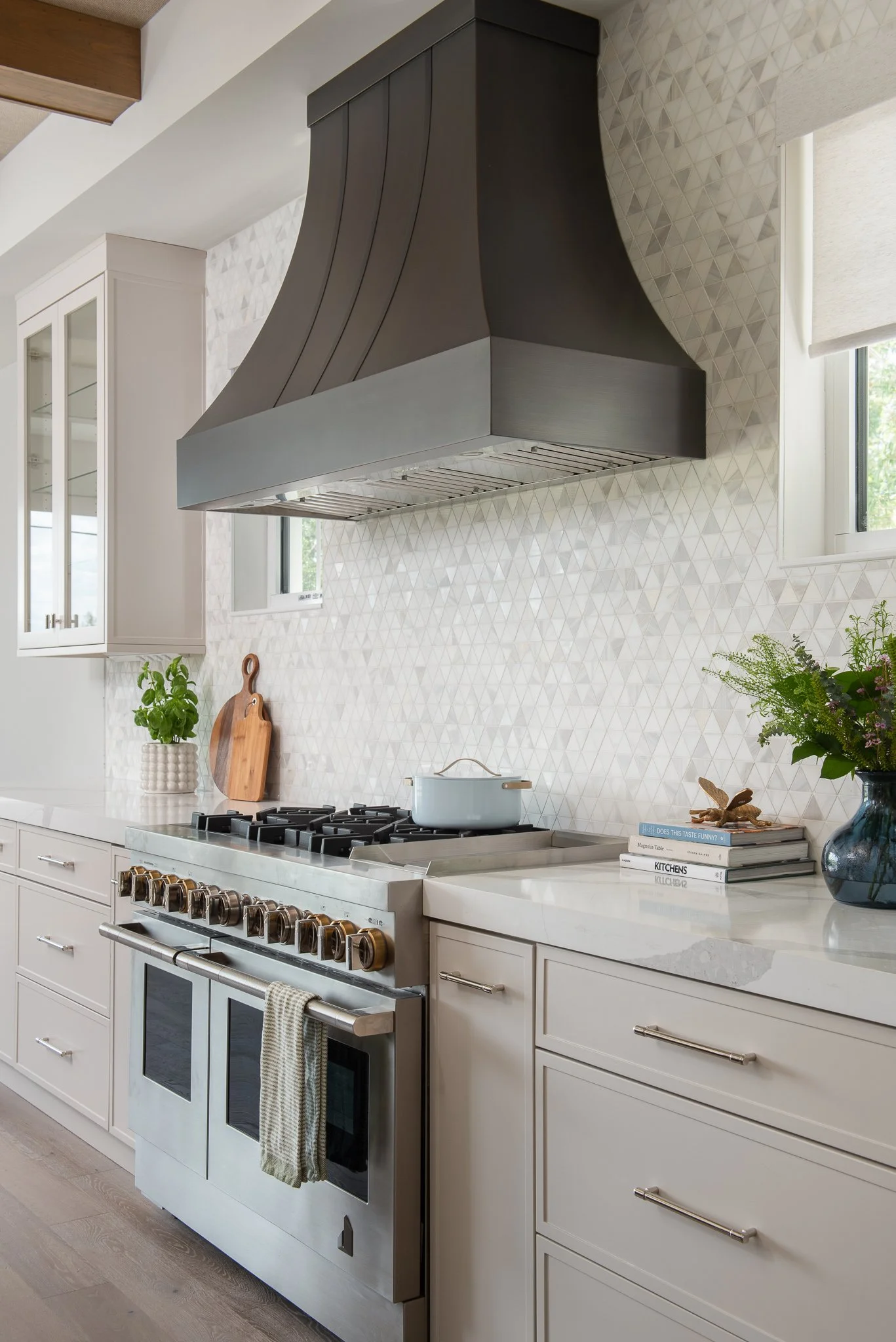 Modern kitchen with white cabinets, stainless steel oven, a white pot on the stove, black range hood, and decorative items including a plant, cutting board, and books on the counter.
