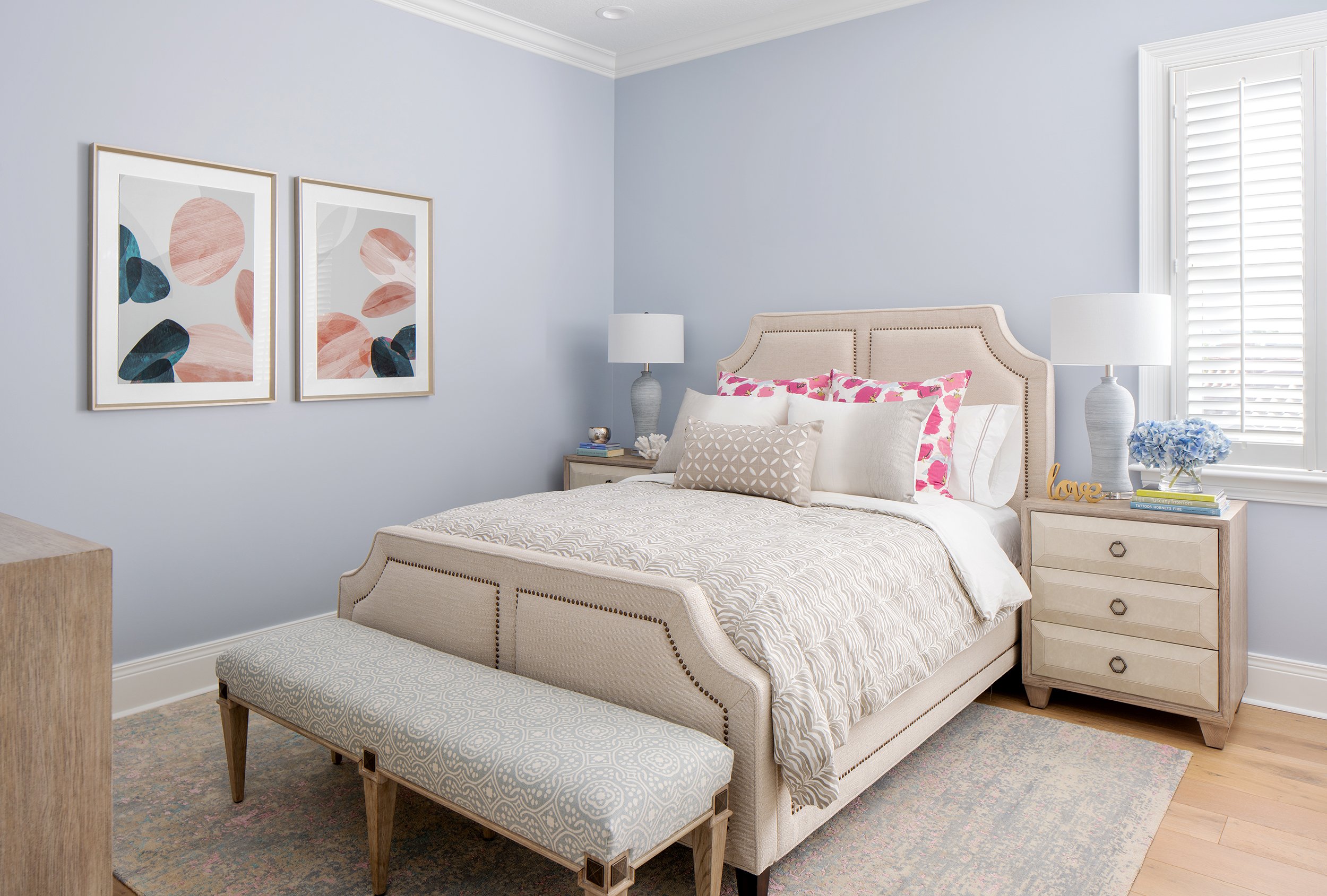 Cozy bedroom with beige upholstered bed, pink and white pillows, white side tables with lamps, blue flowers, and abstract artwork on light blue walls.