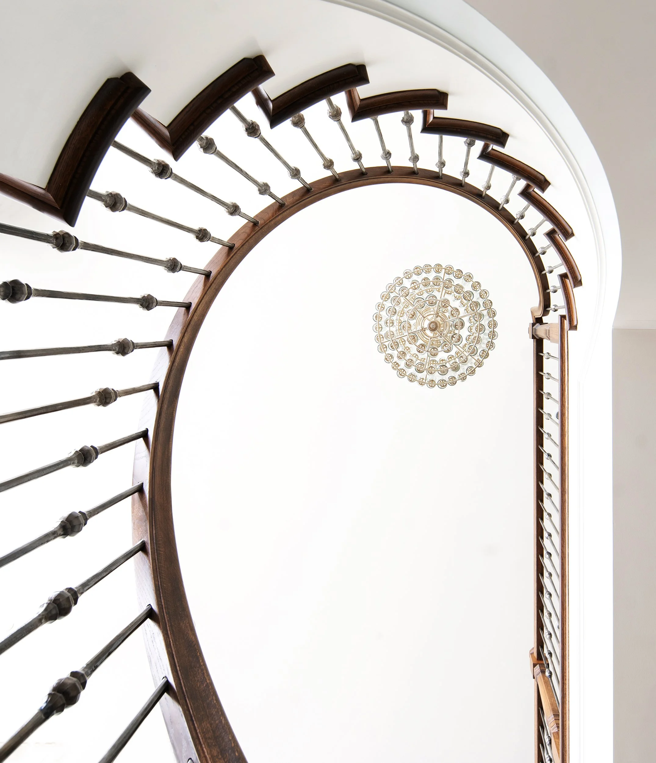 Looking up at a spiral staircase with wooden handrails and metal balusters, with a chandelier hanging from the ceiling at the center.