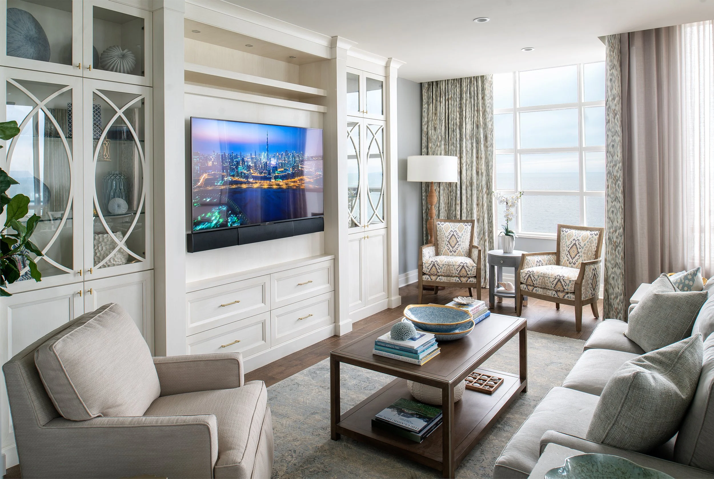 Cozy living room with beige furniture, a wooden coffee table, built-in white shelving, a flat-screen TV, large windows with curtains showing ocean view, and decorative items.