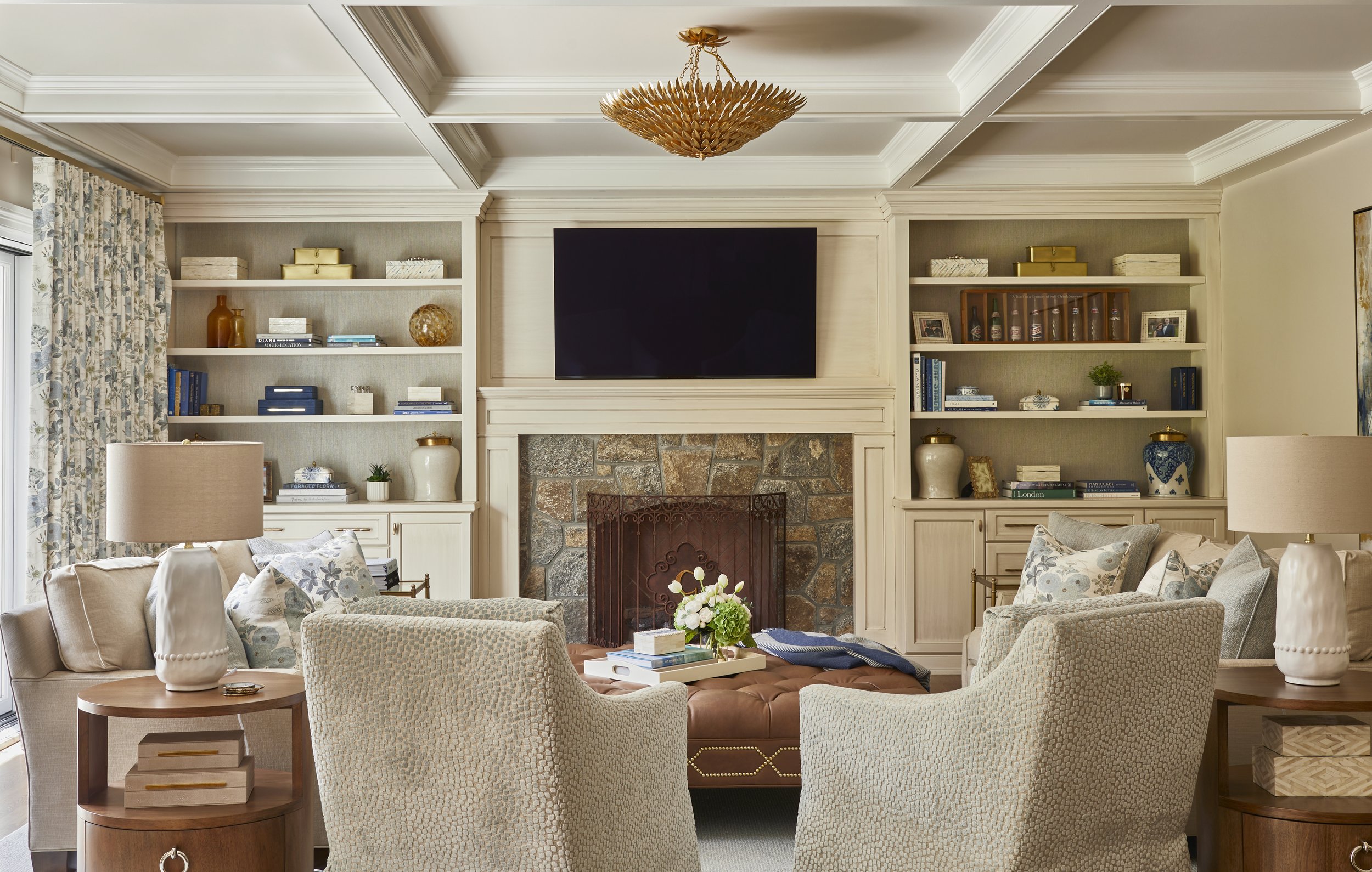 Living room with a stone fireplace, wall-mounted TV, built-in bookshelves, beige couches, armchairs, side tables with lamps, and decorative objects including flowers and books.