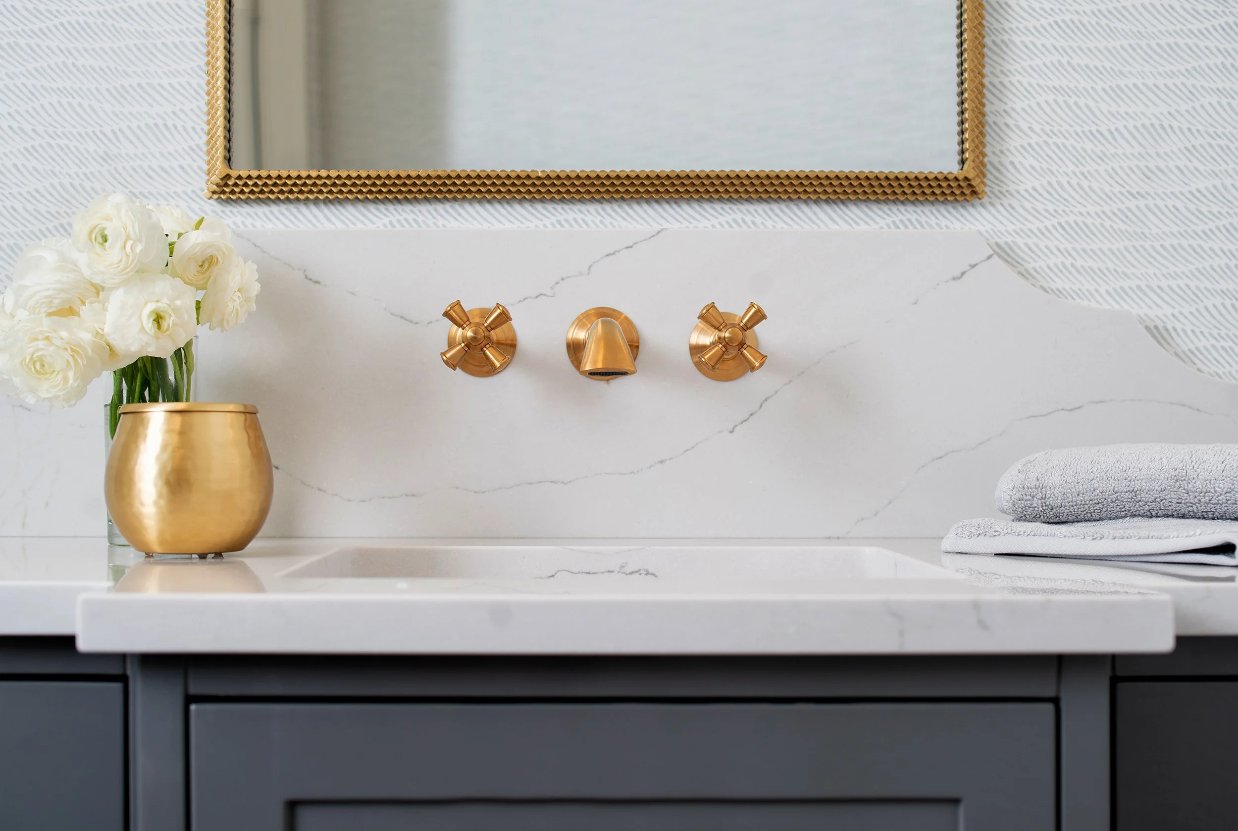 A bathroom vanity with a white marble countertop, a gold vase with white flowers, a gold faucet set, a mirror, and folded towels to the right.