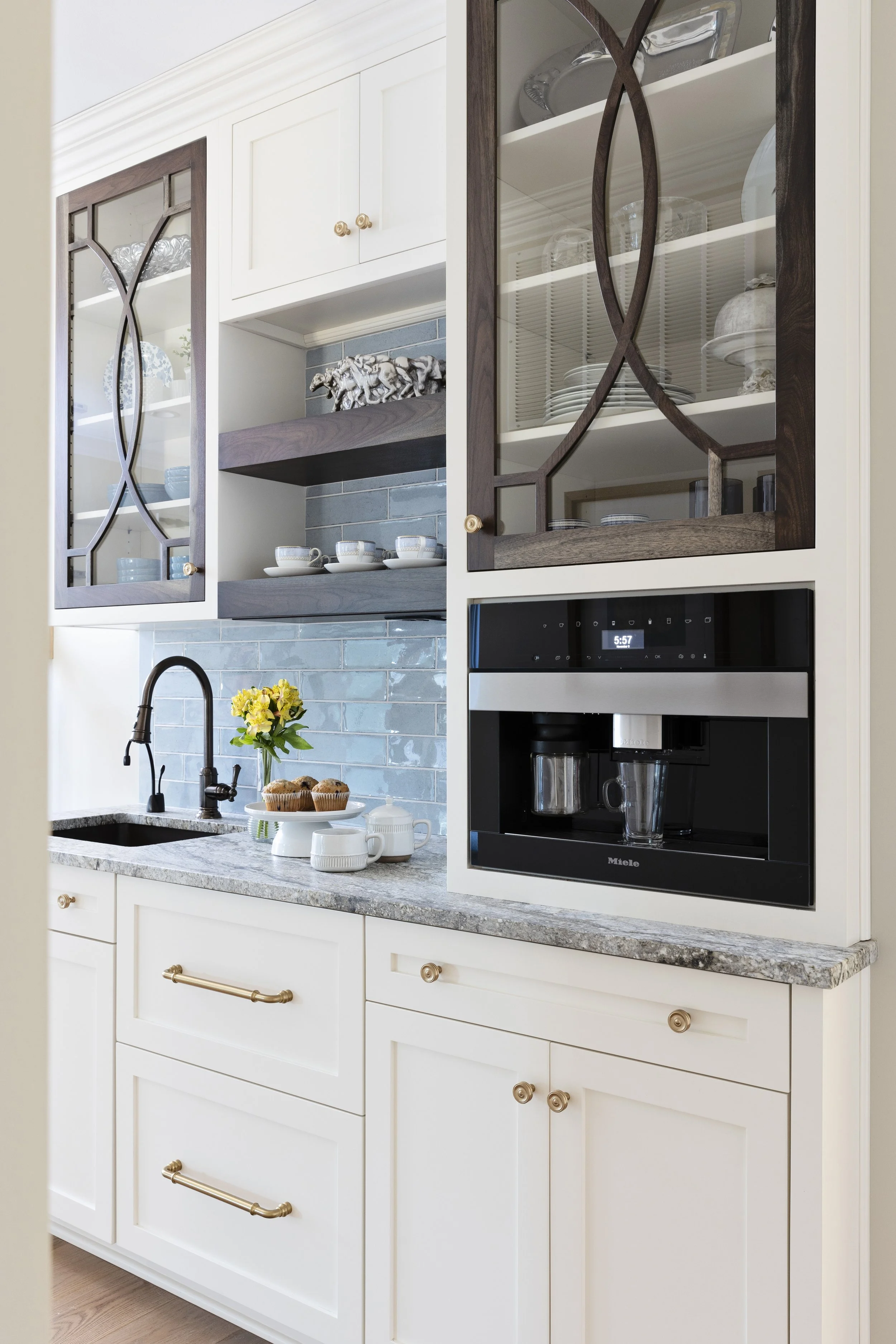 A kitchen with white cabinets, a granite countertop, a black faucet, and a blue tile backsplash. Open glass-front cabinets display dishes and glassware. On the counter, there are a vase with yellow flowers, a plate of muffins, and several cups. A bui
