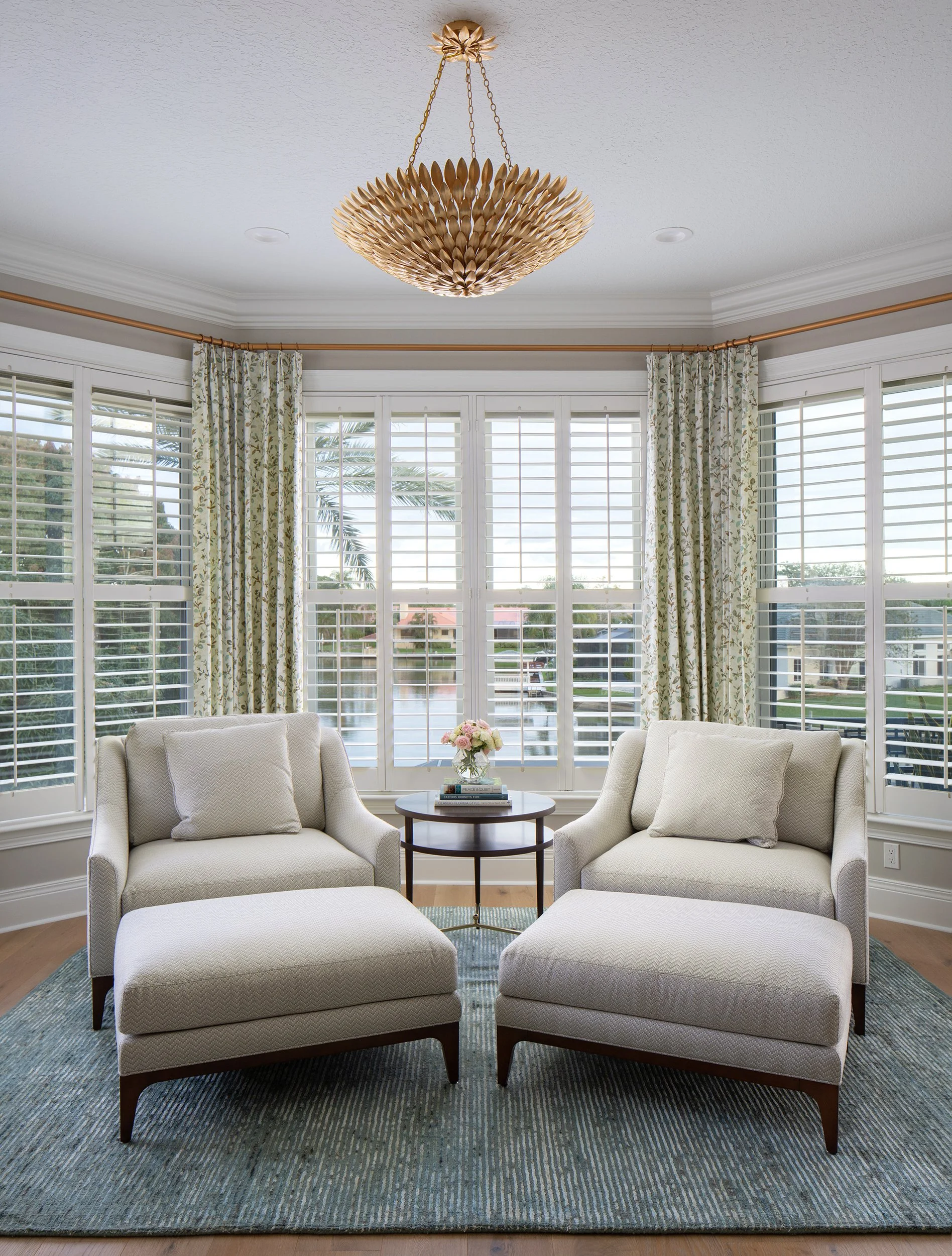 Living room with two white armchairs and a matching ottoman, a small round wooden table with a vase of flowers, large windows with white blinds and patterned curtains, a gold chandelier hanging from the ceiling, and a blue area rug.