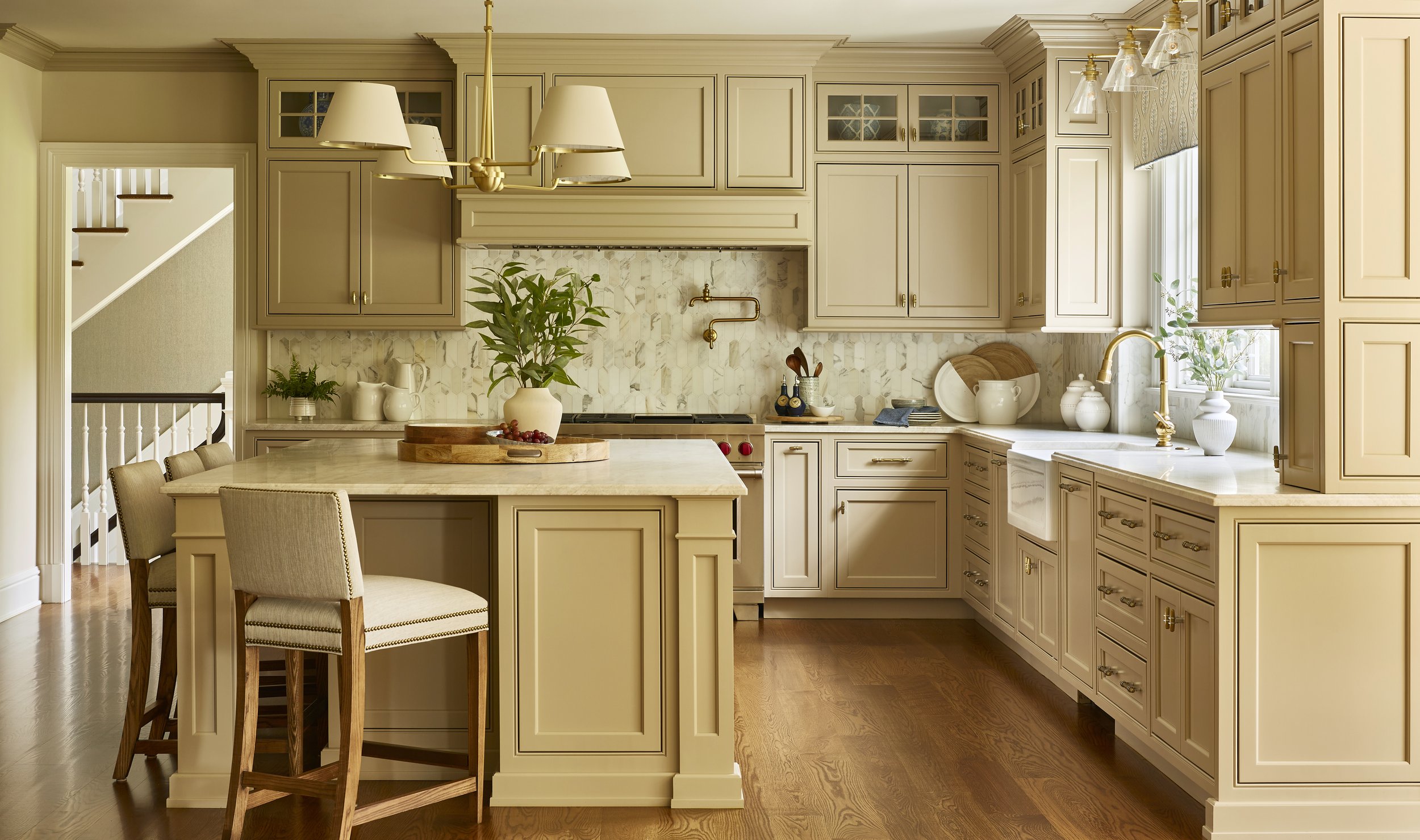 Bright kitchen with beige cabinets, marble backsplash, wooden floors, a kitchen island with beige chairs, and decorative plants near a window.