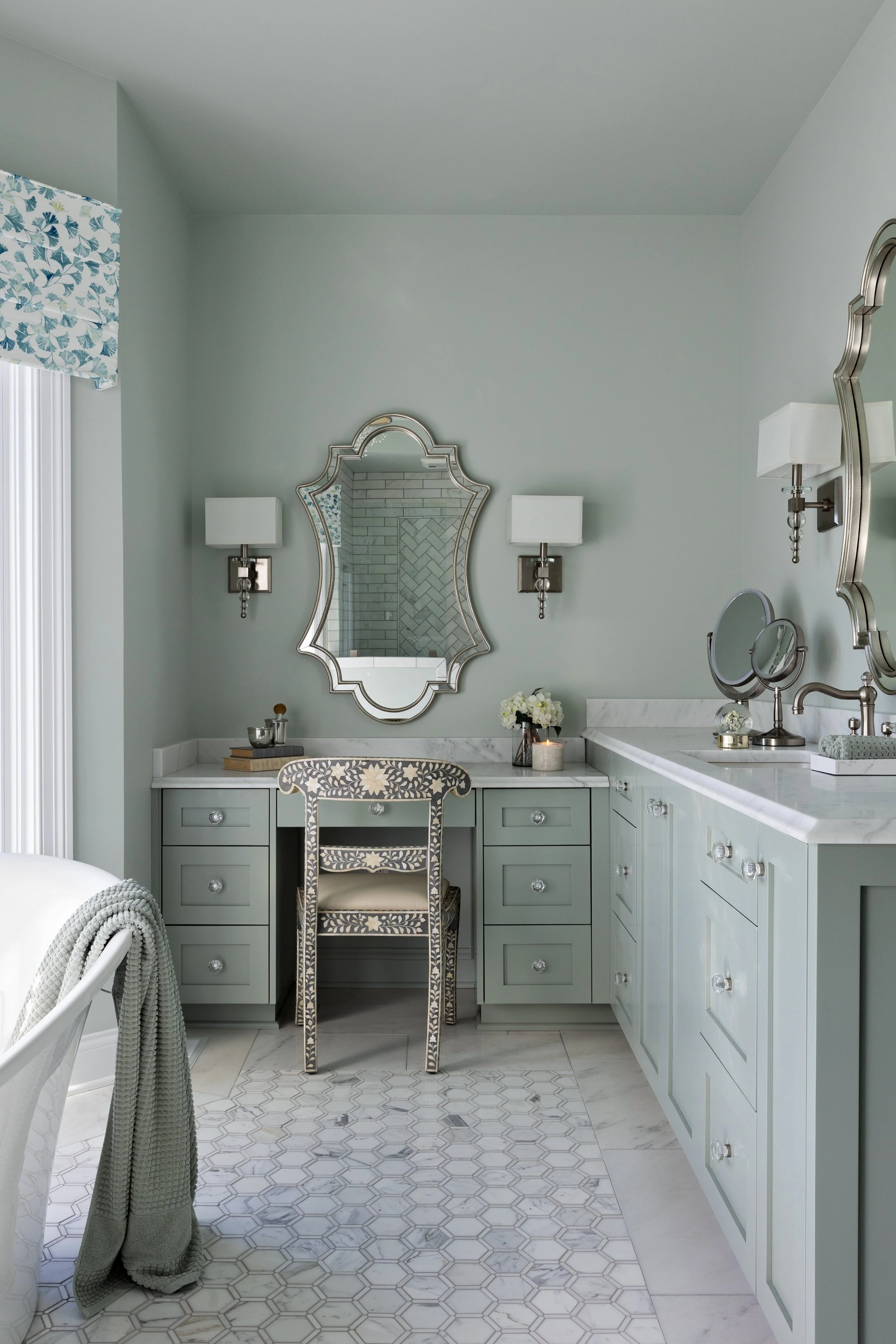 A light green bathroom vanity with a marble countertop, a decorative mirror, wall sconces, a chair with a floral pattern, and various toiletries and decor.