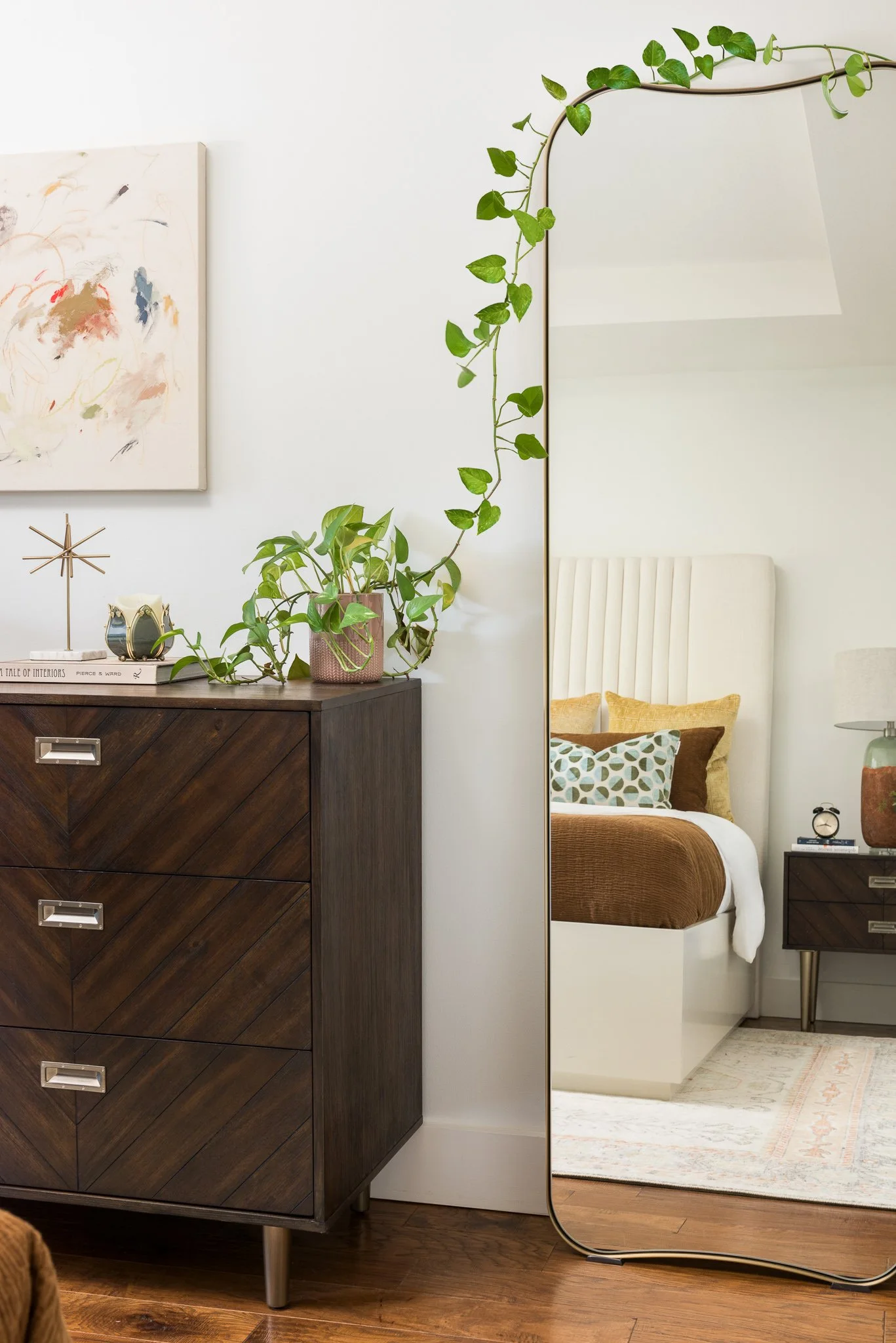 A bedroom with a wooden dresser with a potted plant and decorative items on top, a mirror with a trailing vine, a bed with yellow and patterned pillows, and a nightstand with a lamp and clock.