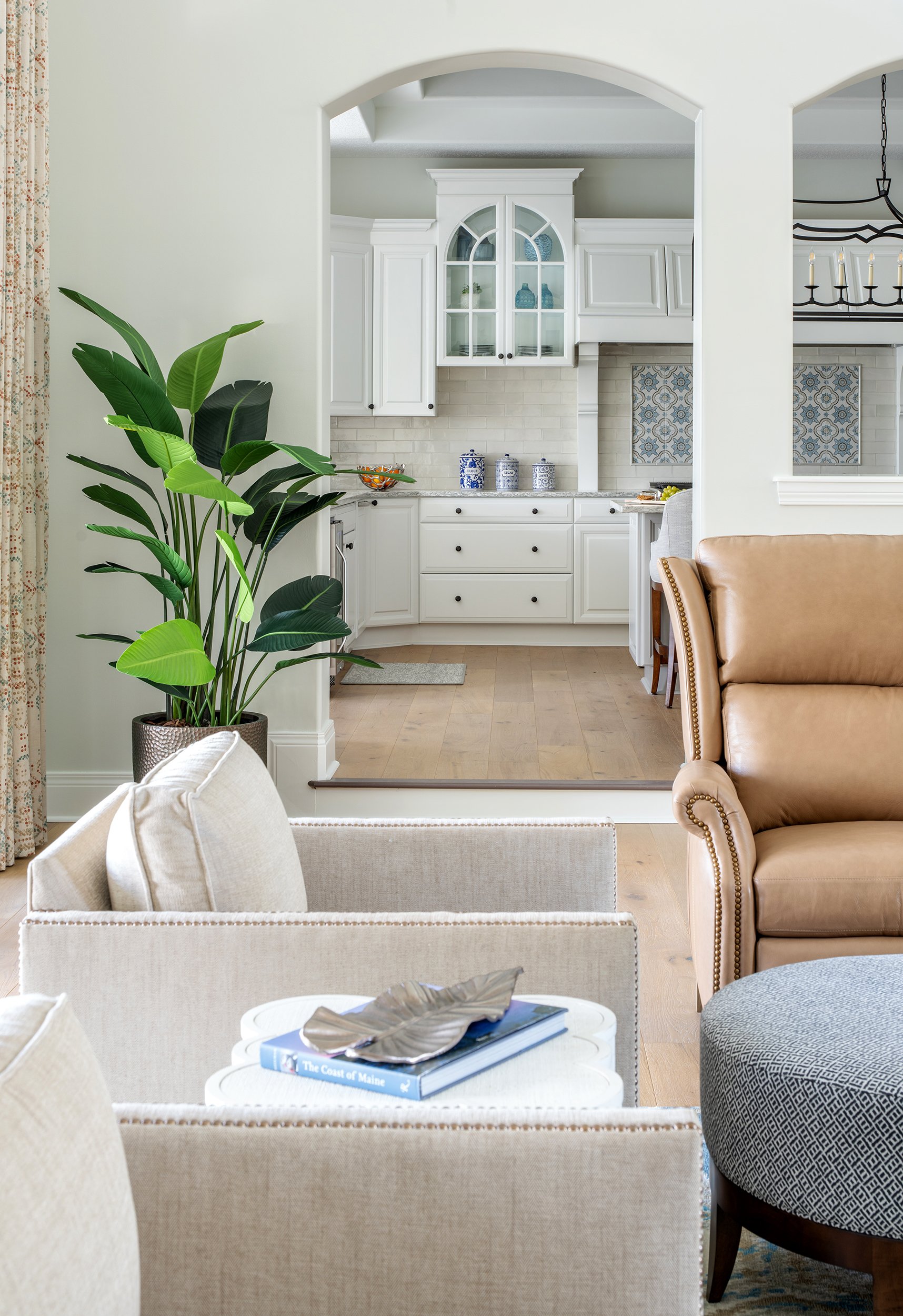 View of a bright living room with beige and tan furniture, a large green potted plant, and a kitchen with white cabinets and light wood flooring in the background.