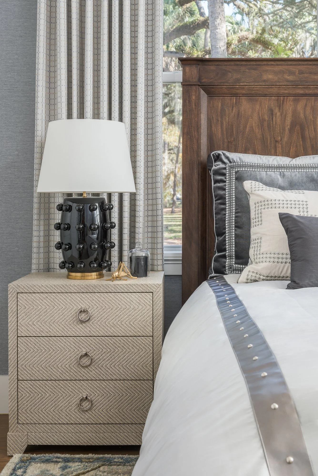A bedroom nightstand with a black ceramic lamp with knobs, a glass cloche, and small decorative items, next to a bed with a wooden headboard, pillows, and a window with trees outside.