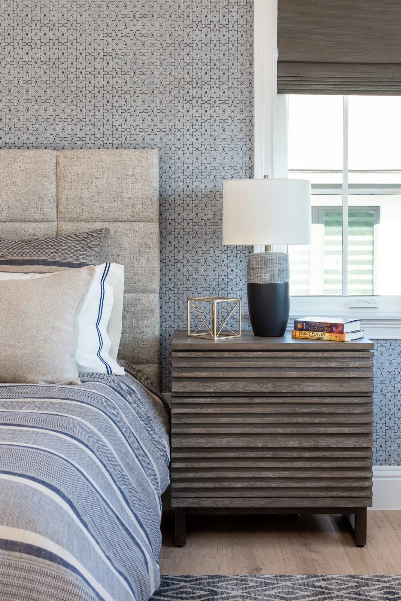 A bedroom corner featuring a bed with striped bedding, beige pillows, and a gray upholstered headboard. Beside the bed, a wooden nightstand holds a modern lamp with a black and gray base, a decorative gold geometric sculpture, and a small stack of bo