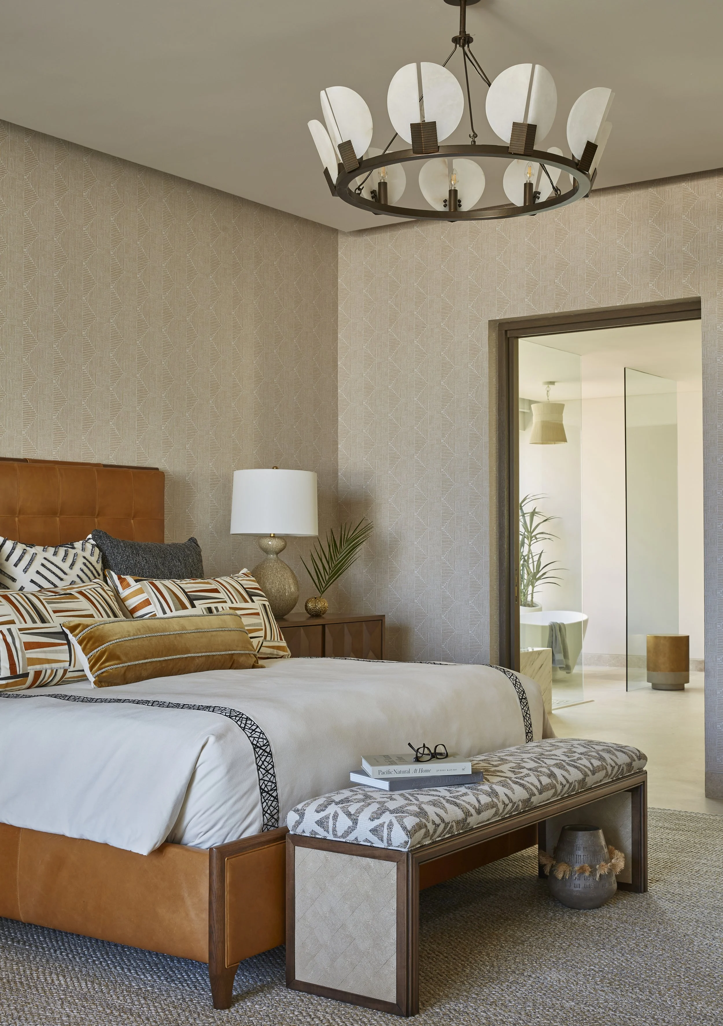 A cozy bedroom with a tan upholstered bed, patterned pillows, and a wooden bench at the foot of the bed. A bedside table with a white lamp and decorative items, and an open doorway leading to a bathroom with a bathtub, plant, and bathroom accessories