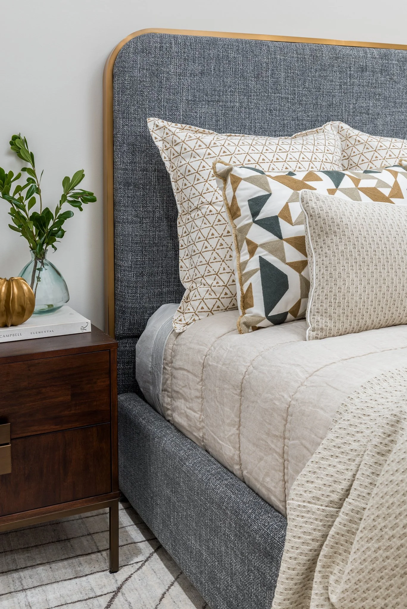 Close-up of a bed with a dark gray upholstered headboard, beige bedding, and decorative pillows with geometric patterns, next to a wooden nightstand with a glass vase of green leaves and a gold decorative object.