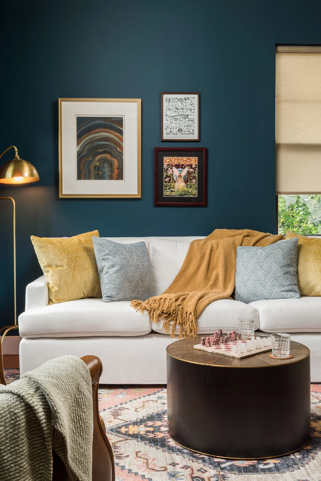Living room with a white sofa, yellow and gray pillows, a brown throw blanket, a round wooden coffee table with chess and drinks, a dark teal wall with framed artwork, a window with beige shades, and a gold floor lamp.