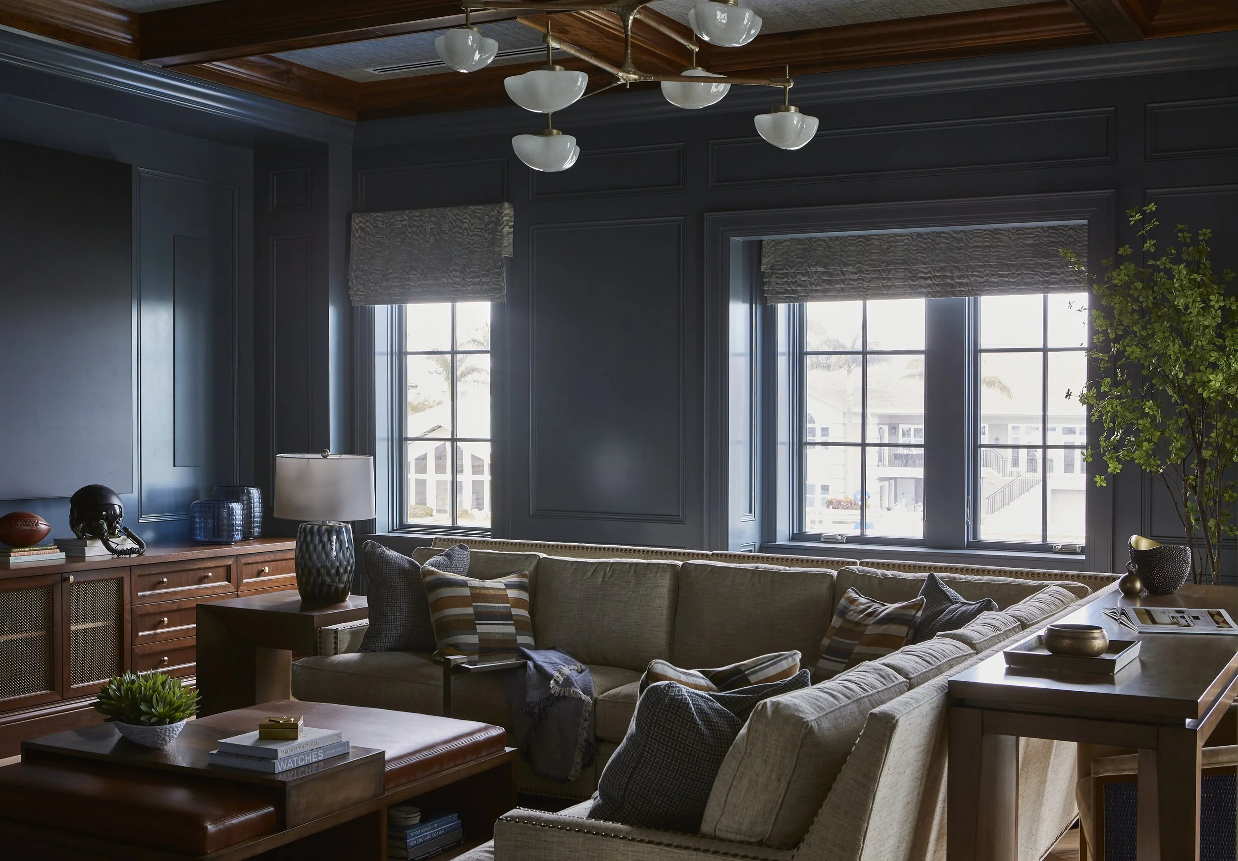 Interior living room with dark blue paneled walls, large windows, beige sectional sofa with patterned pillows, wooden coffee table with books and a plant, side tables with lamps, and a wooden credenza with decorative items, including a football and a