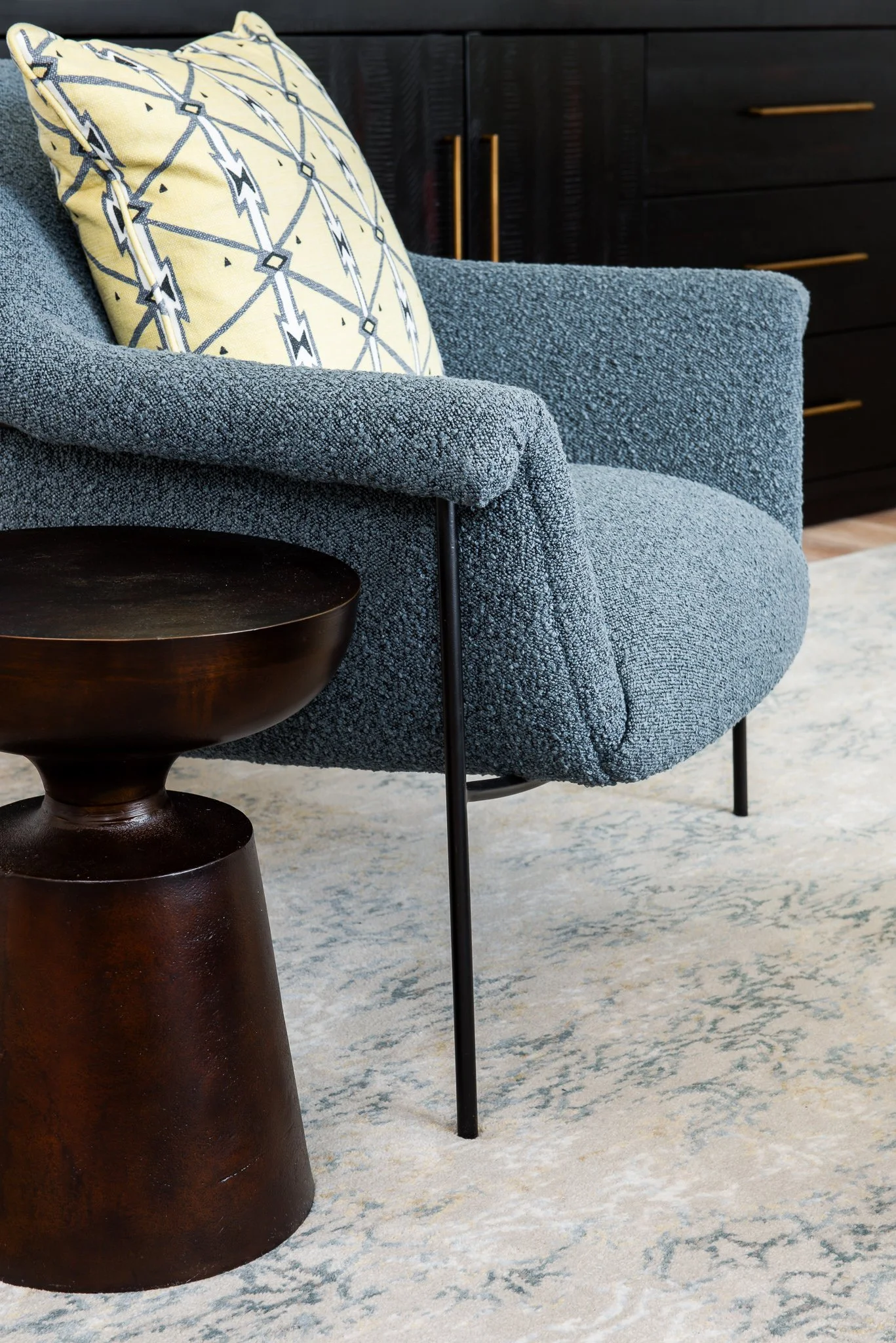 Close-up of a blue textured armchair with a yellow and black patterned cushion, next to a dark brown wooden side table, on a light-colored patterned rug.